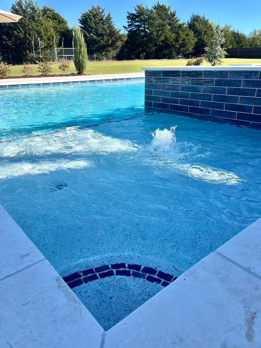 Close-up of a backyard pool with blue water, a built-in hot tub, and a brick wall feature. In the background, there is a grassy area with trees and a fenced yard under a clear blue sky.