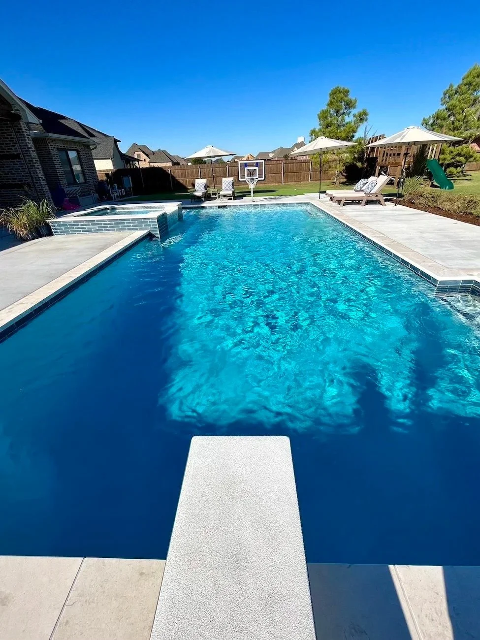 A backyard swimming pool with a diving board, surrounded by a concrete deck, poolside chairs, umbrellas, and a small play slide, with a wooden fence and houses in the background under a clear blue sky.