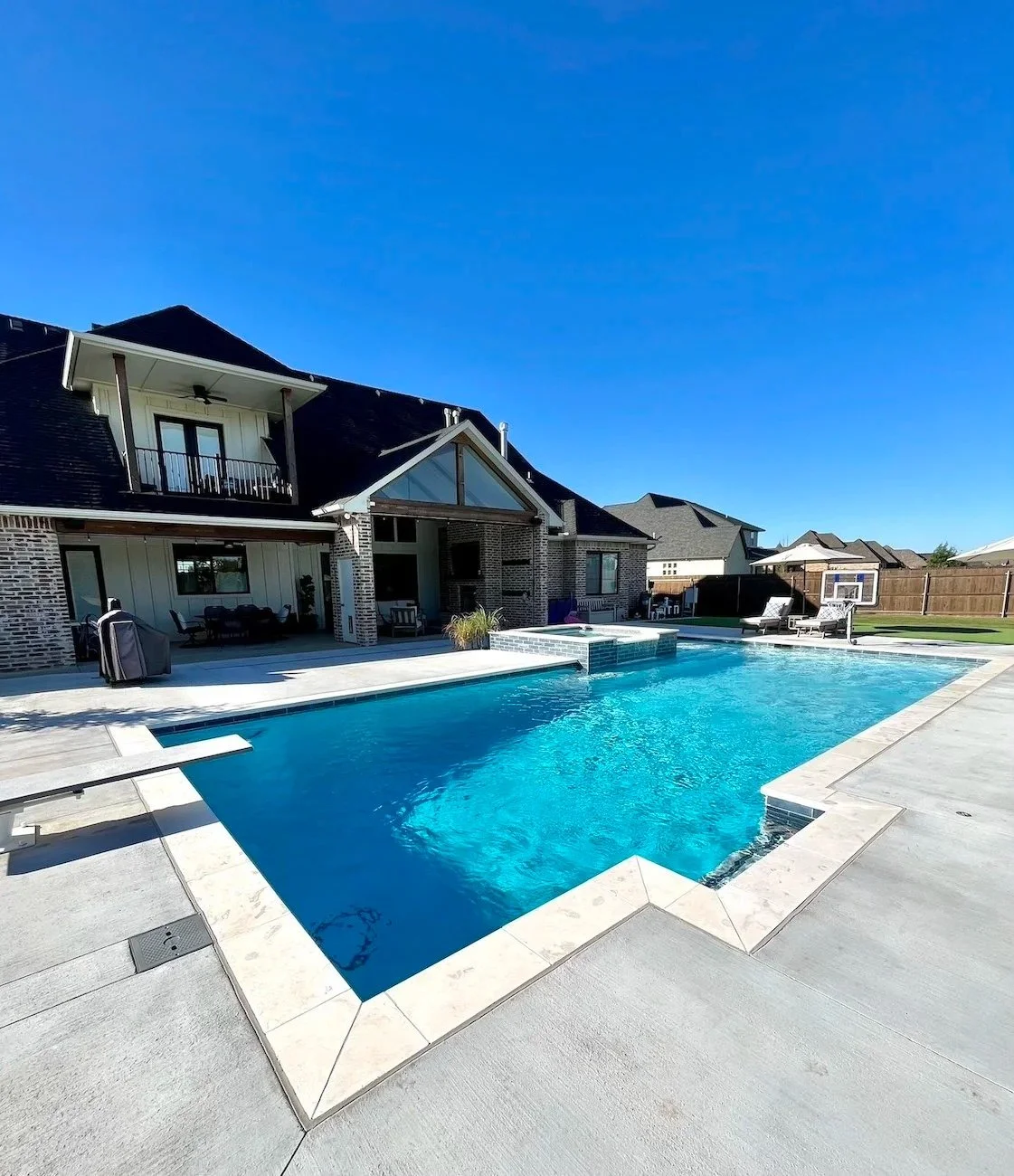 Backyard with a modern house, swimming pool, patio furniture, and a basketball hoop under a clear blue sky.