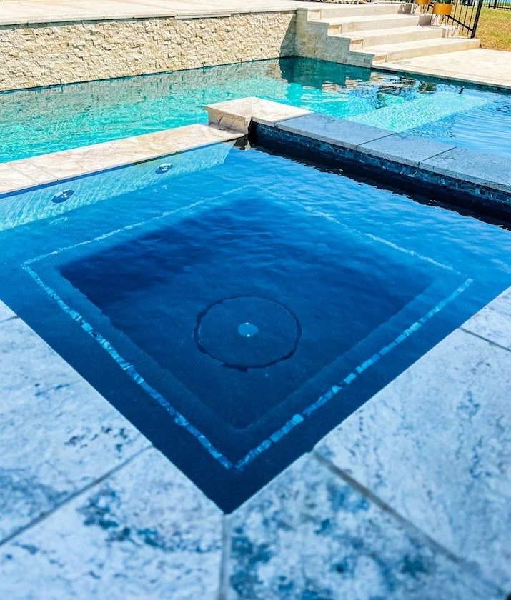 A close-up view of an empty hot tub with clear water, tiled surface, and adjacent swimming pool with steps leading into it.