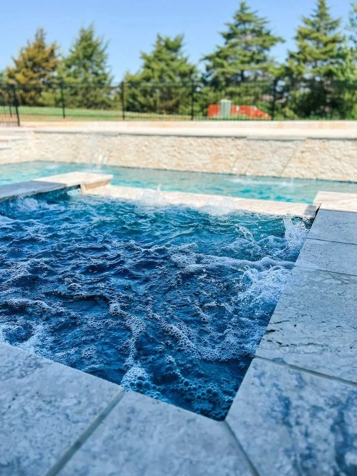 A swimming pool with bubbling water, surrounded by stone tiles, with trees and a fence in the background under a clear blue sky.
