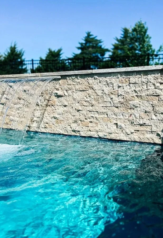 Close-up of a waterfall spilling into a pool of blue water from a stone wall, with trees and a metal fence in the background.