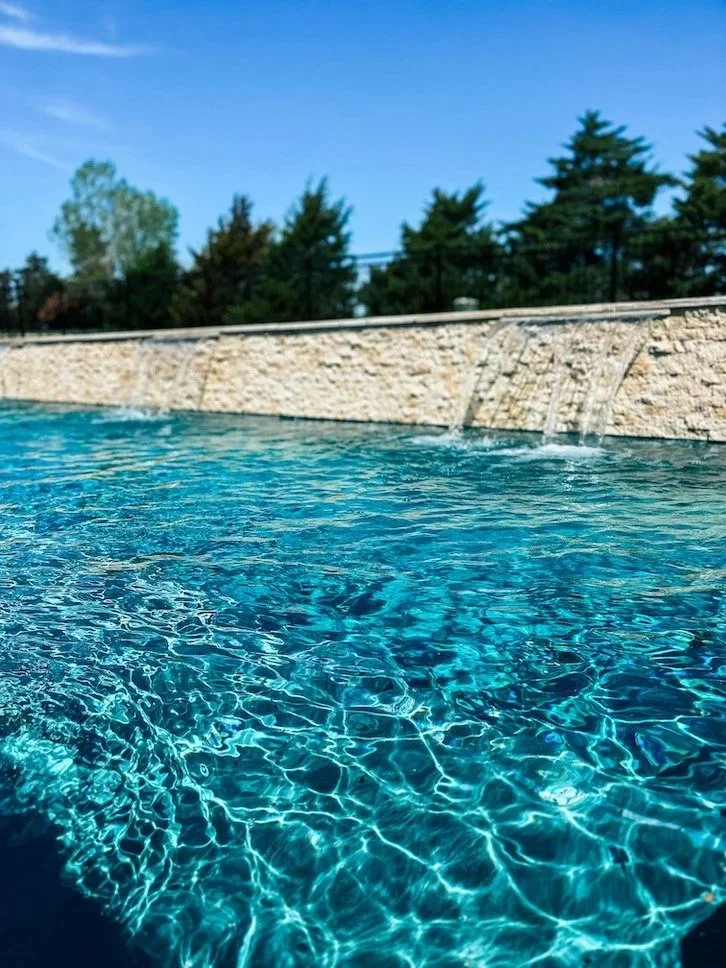 A close-up of a swimming pool with clear blue water, a stone waterfall feature on the side, and trees in the background under a blue sky.
