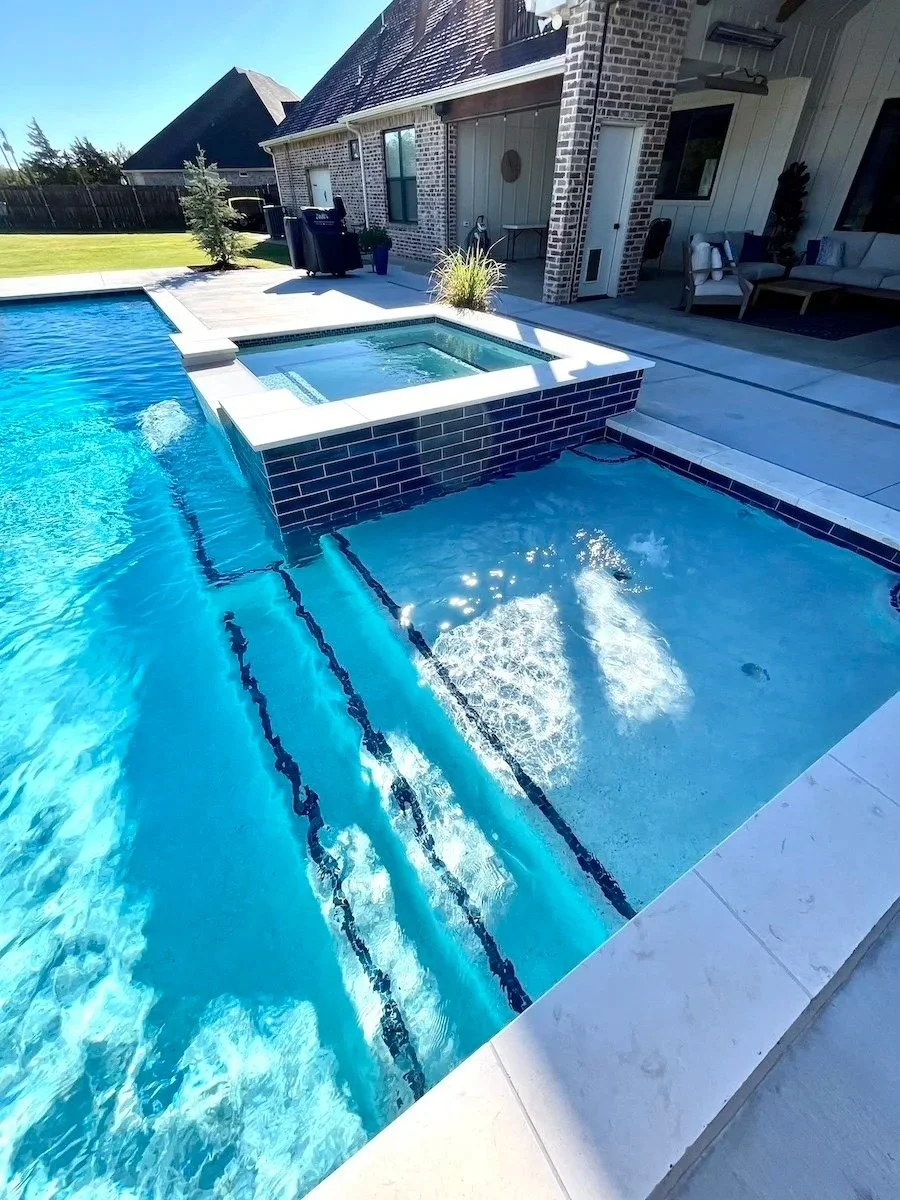 A backyard swimming pool with a hot tub and seating area, surrounded by a patio, with a house in the background.