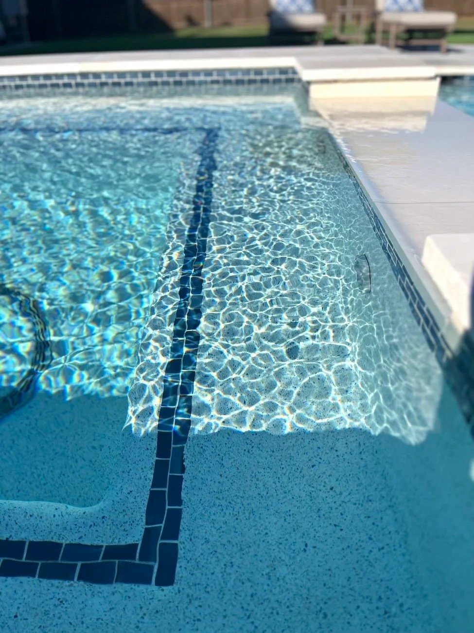A swimming pool with clear, blue water and a submerged ladder with a black edge, surrounded by a white tiled deck, in a sunny outdoor setting.