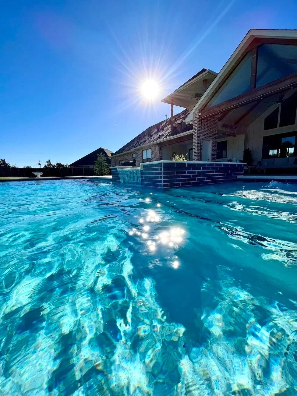 Residential backyard swimming pool with clear blue water, a house with a sloped roof and large windows, and the sun shining brightly in a clear blue sky.