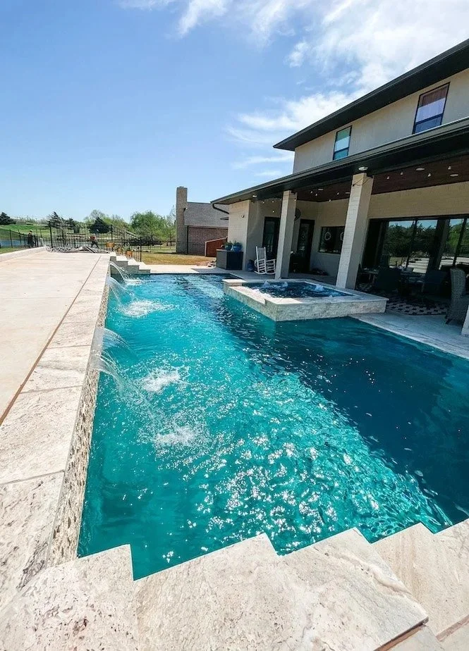 Outdoor swimming pool with water features, adjacent to a house with a covered patio, under a partly cloudy sky.