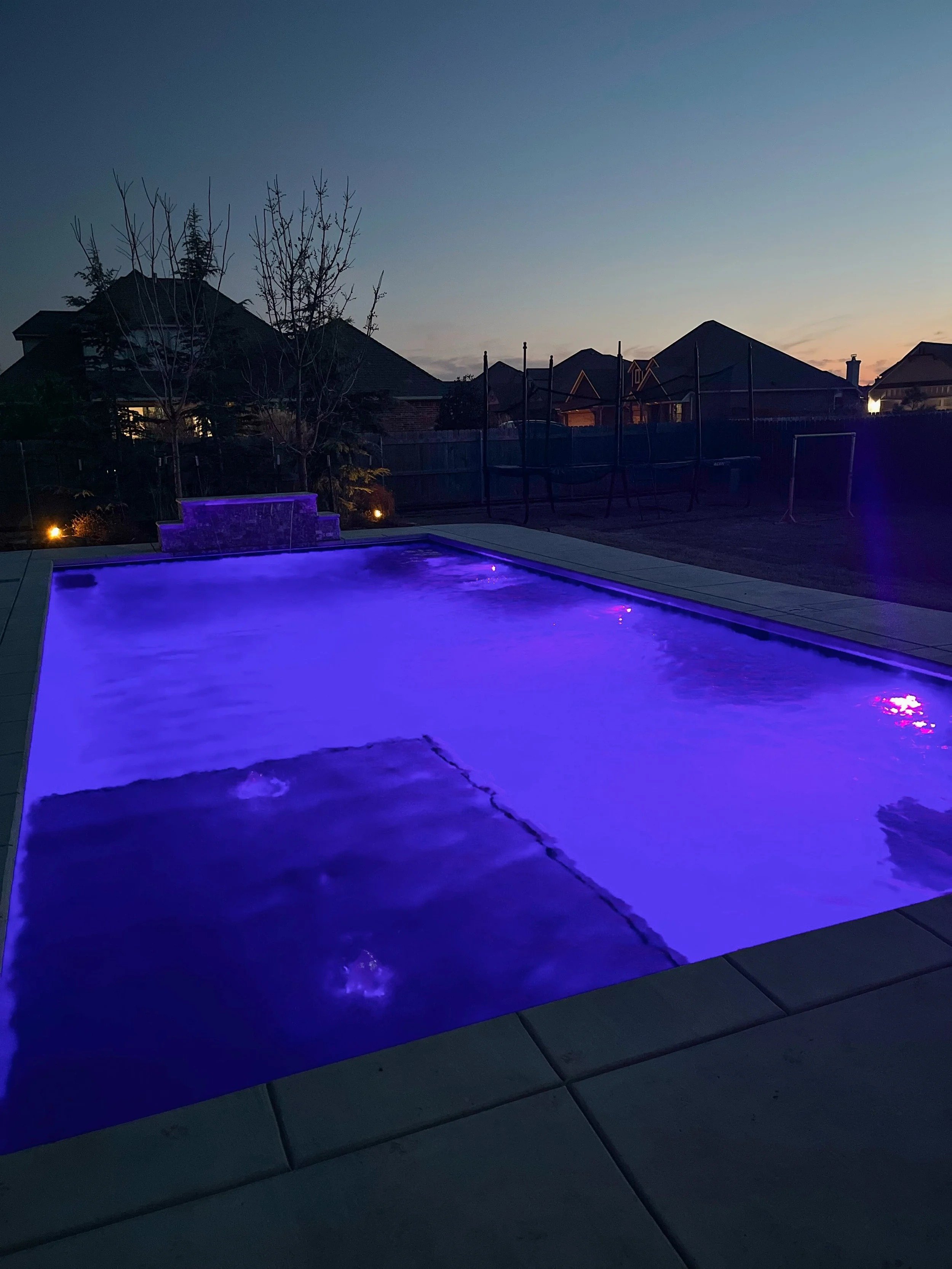 A backyard swimming pool illuminated with purple lighting at sunset, with trees and houses visible in the background.