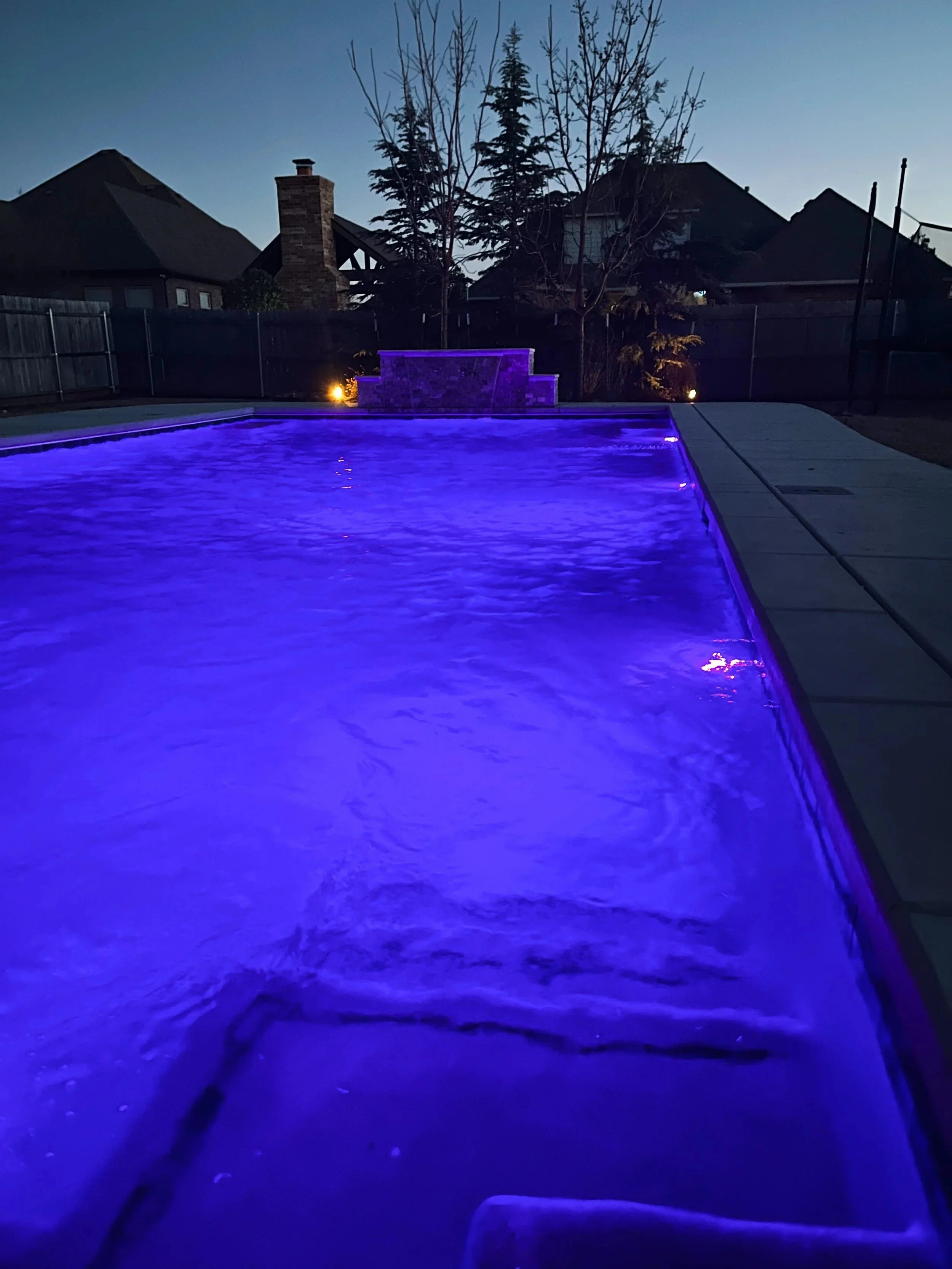 Nighttime backyard with an illuminated pool glowing blue, surrounded by a concrete deck, with trees and houses in the background.