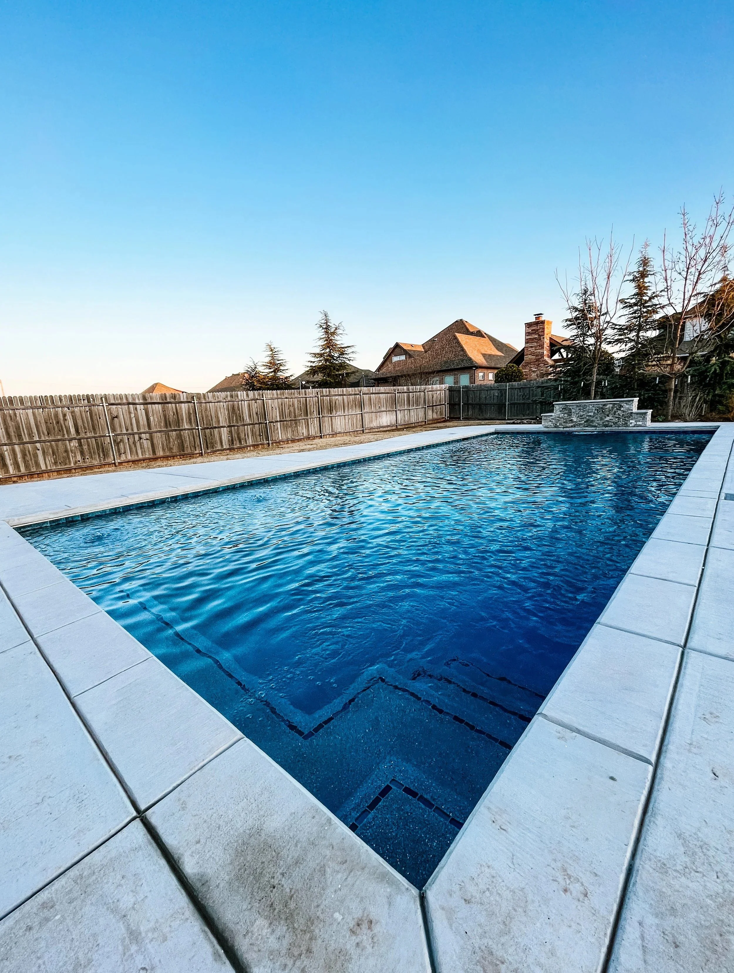 In-ground swimming pool with a stone water feature at one end, surrounded by a concrete deck and a wooden fence, with houses and trees in the background under a clear blue sky.