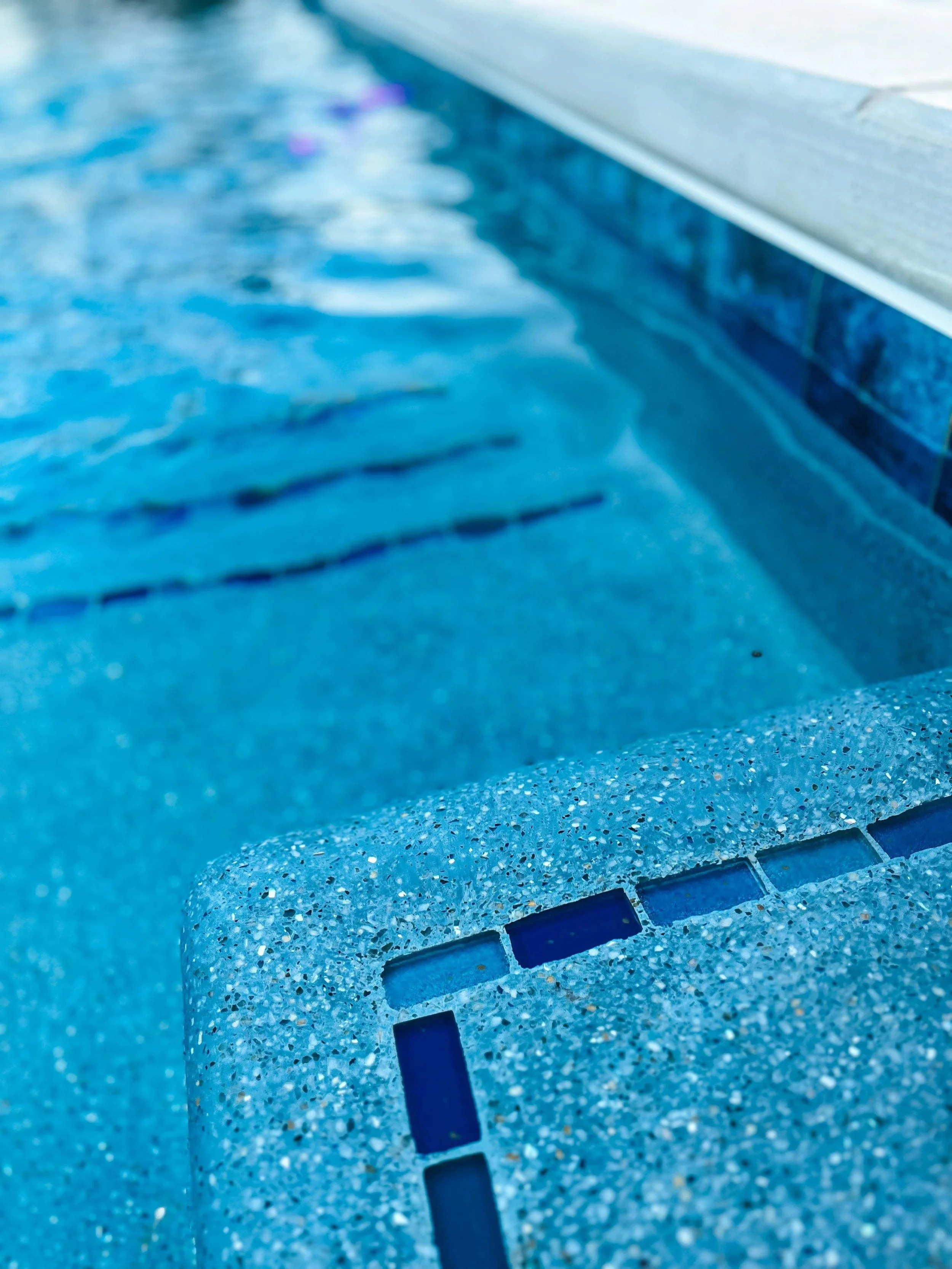 Close-up of the edge of a swimming pool with blue tiles and a blue tiled coping, showing the water surface with light reflections.