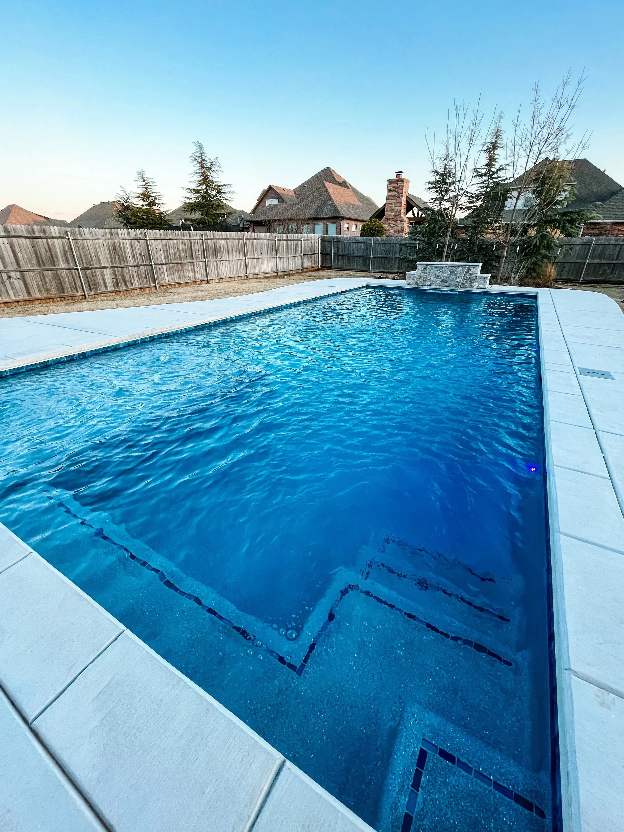 Backyard swimming pool with water feature, surrounded by concrete deck and wooden fence, with houses and trees in the background under a clear sky.