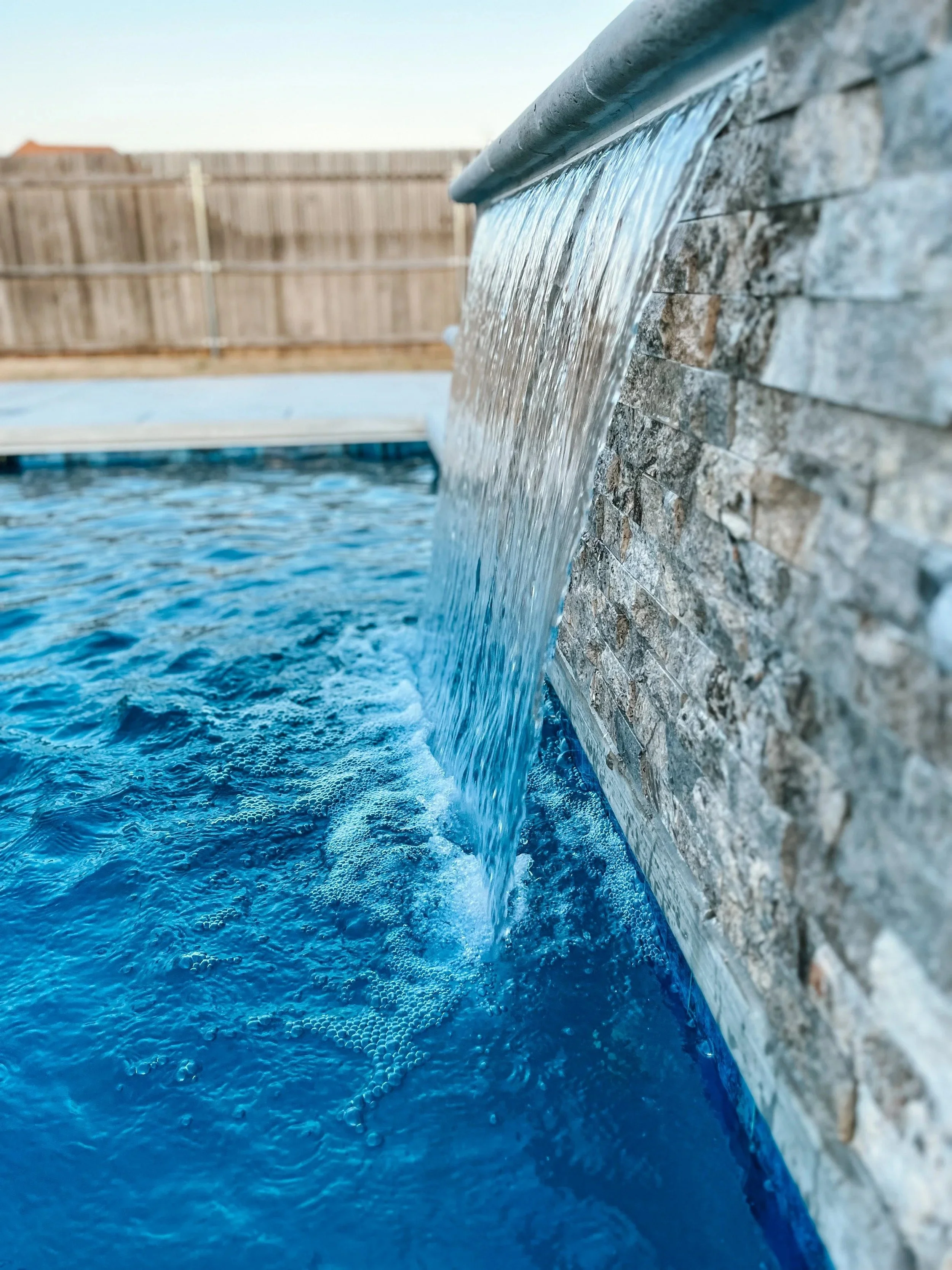 Water is flowing from a spout onto a textured stone wall, with a swimming pool below.