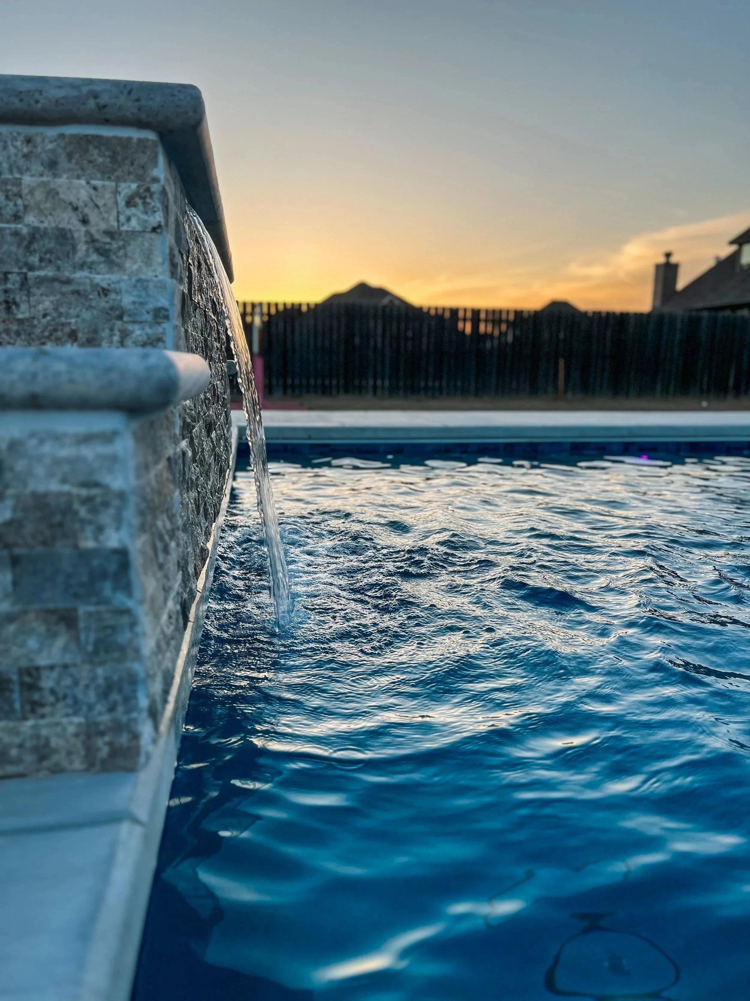 Close-up view of a stone water fountain with water streaming from a spout into a pool, set against a sunset sky with buildings and a wooden fence in the background.