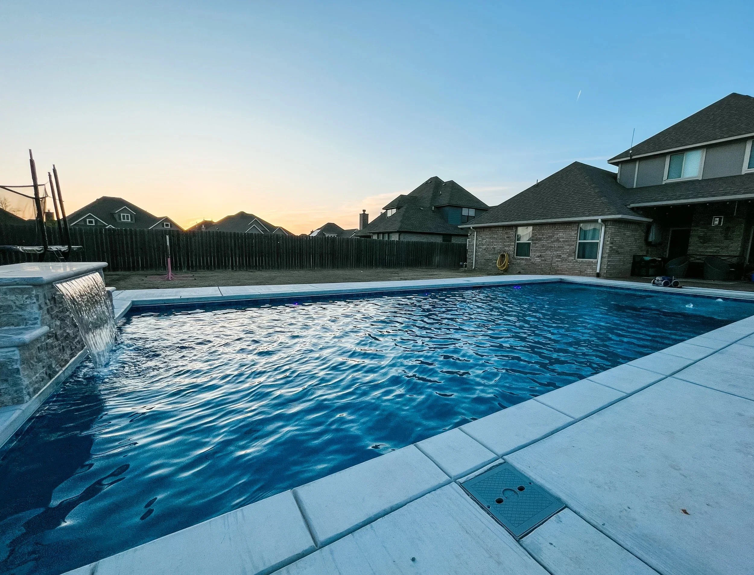 A backyard swimming pool at sunset with houses and a clear sky in the background.