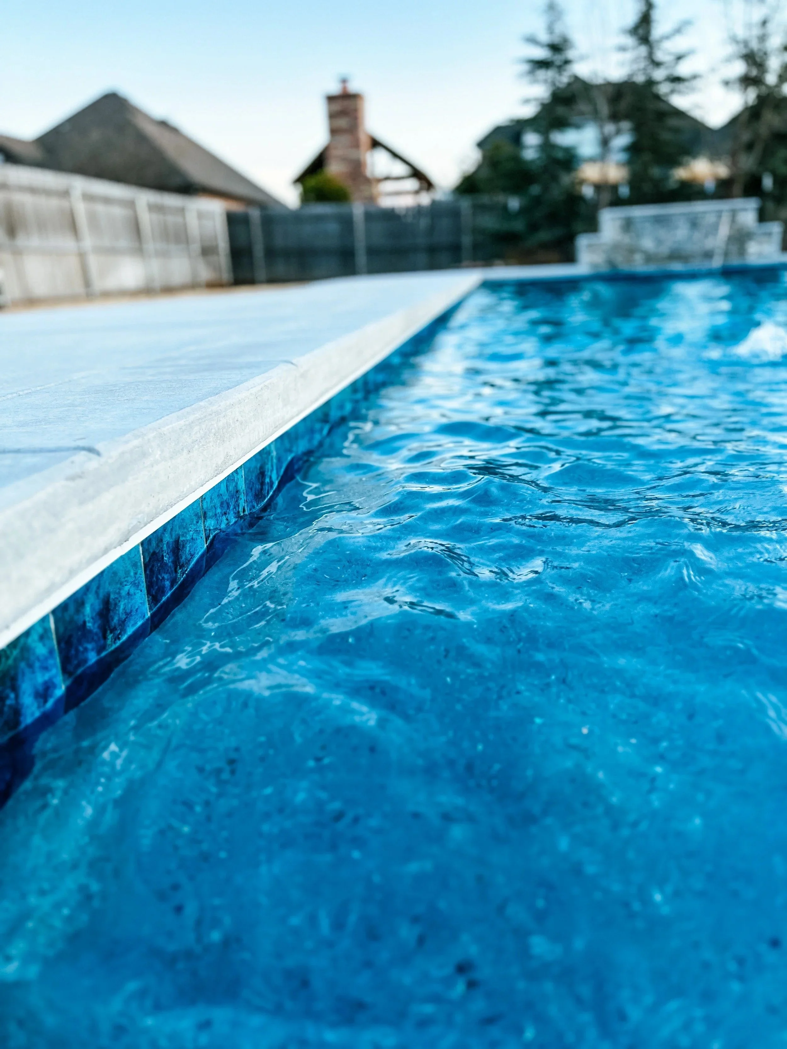 Close-up view of the edge of a swimming pool with blue water, surrounded by a white pool deck, with a blurry background of houses and trees.