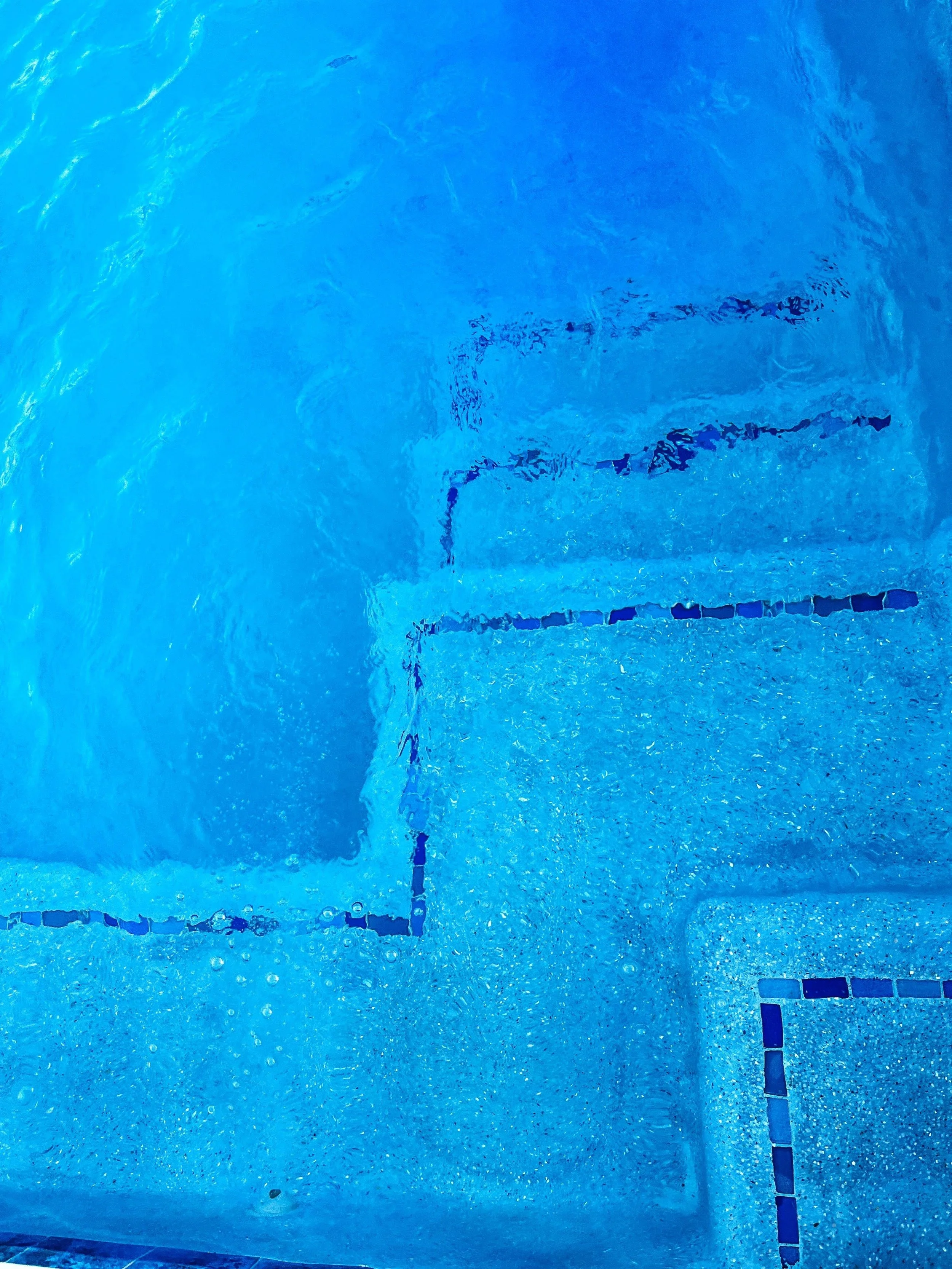 An underwater view of a swimming pool with blue tiles and lane markings, showing ripples and bubbles on the water surface.