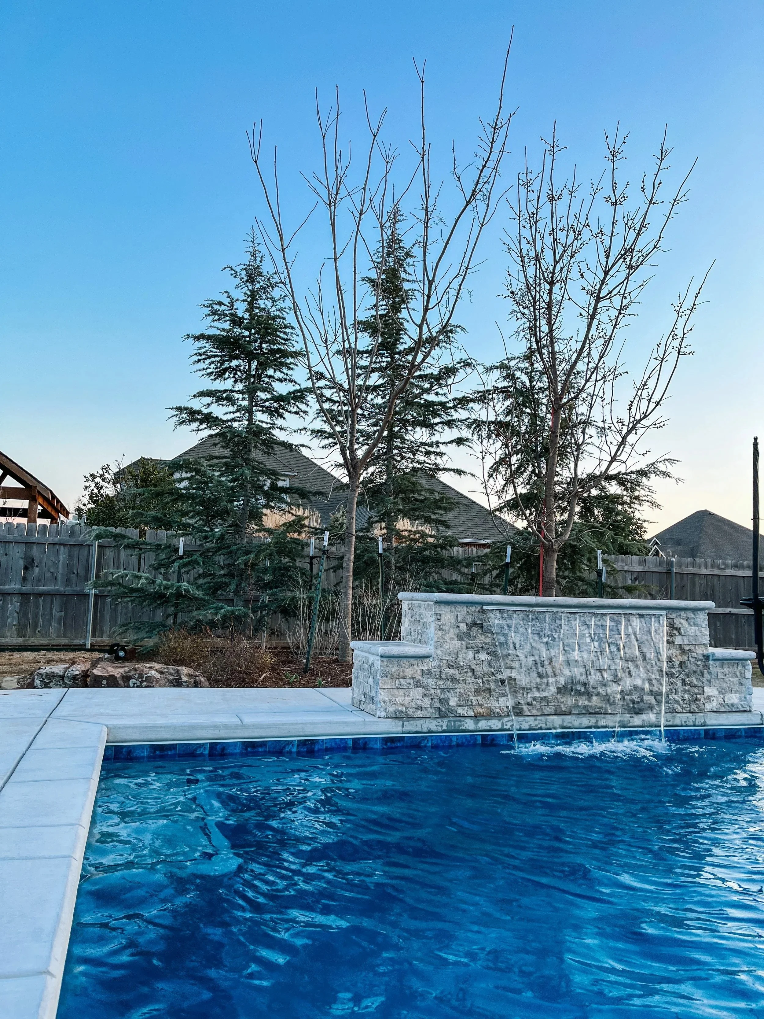 A backyard swimming pool with a stone waterfall feature, surrounded by a concrete deck, with a wooden fence, trees, and houses in the background under a clear blue sky.