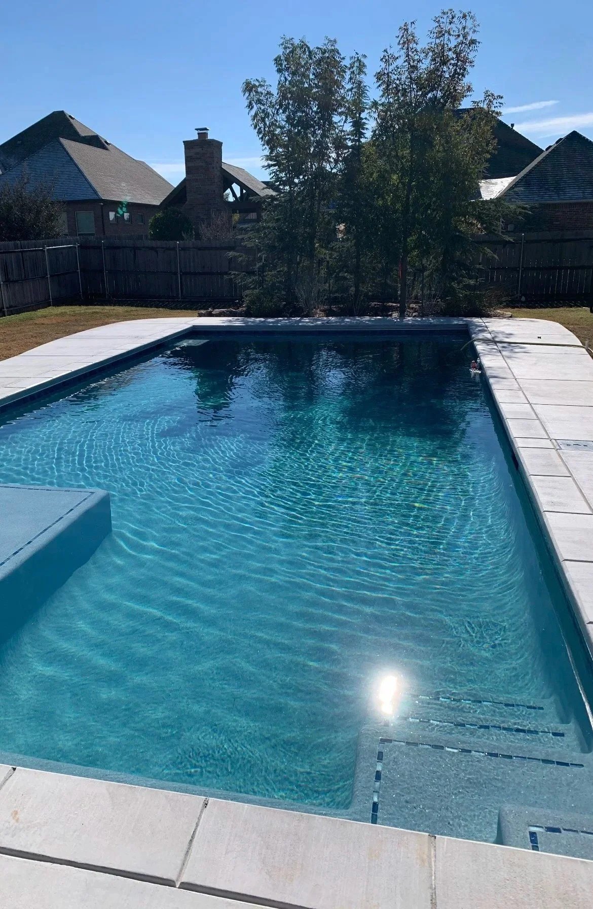A backyard swimming pool with clear blue water, surrounded by a white concrete deck. There are trees and a wooden fence in the background, with houses and a chimney visible beyond the fence. The sky is clear and sunny.