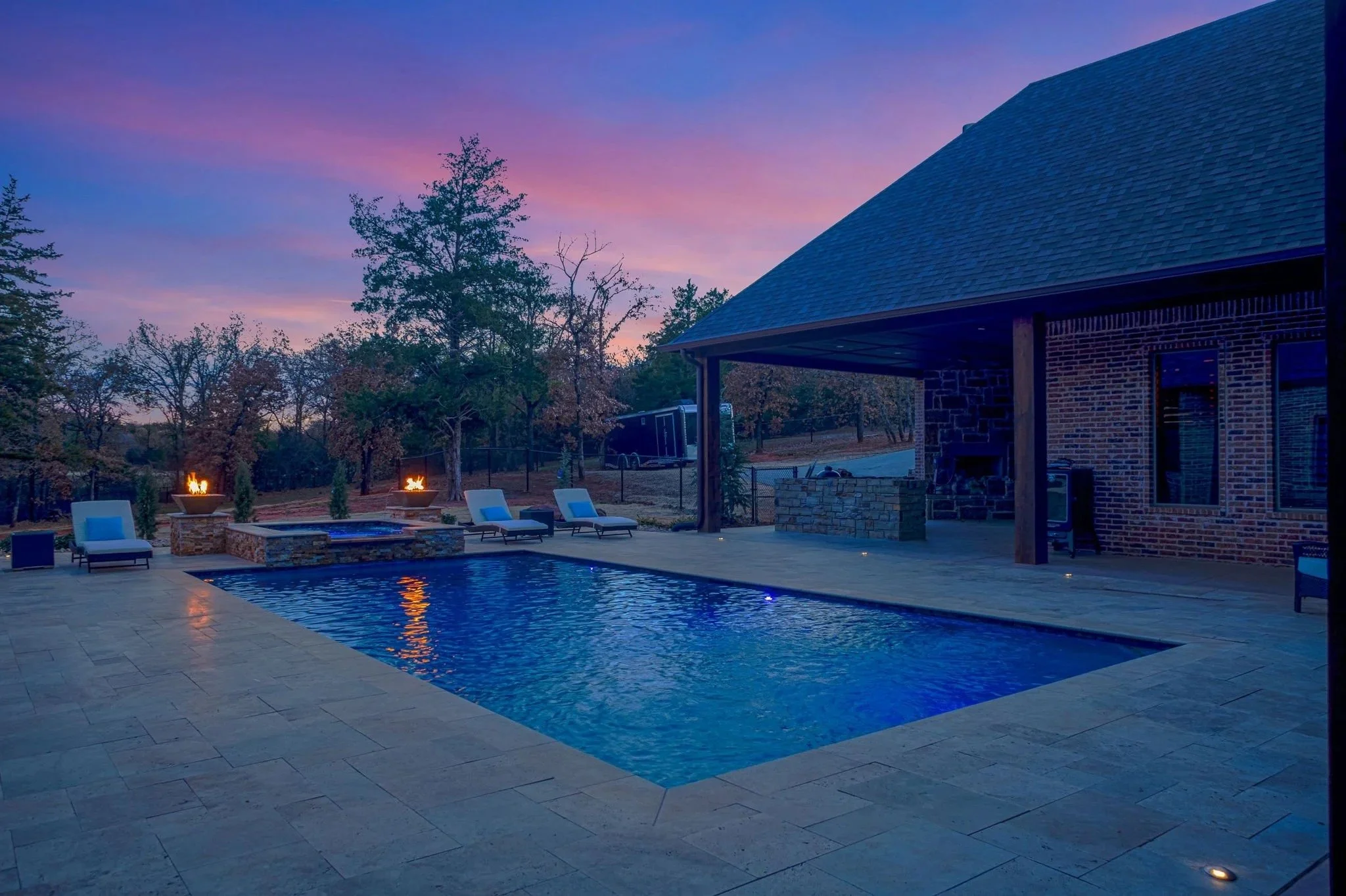 A backyard scene at dusk with a swimming pool illuminated with blue lights, surrounded by lounge chairs and fire pits. There are trees and a brick house with a covered porch in the background, and the sky shows a pink and purple sunset.