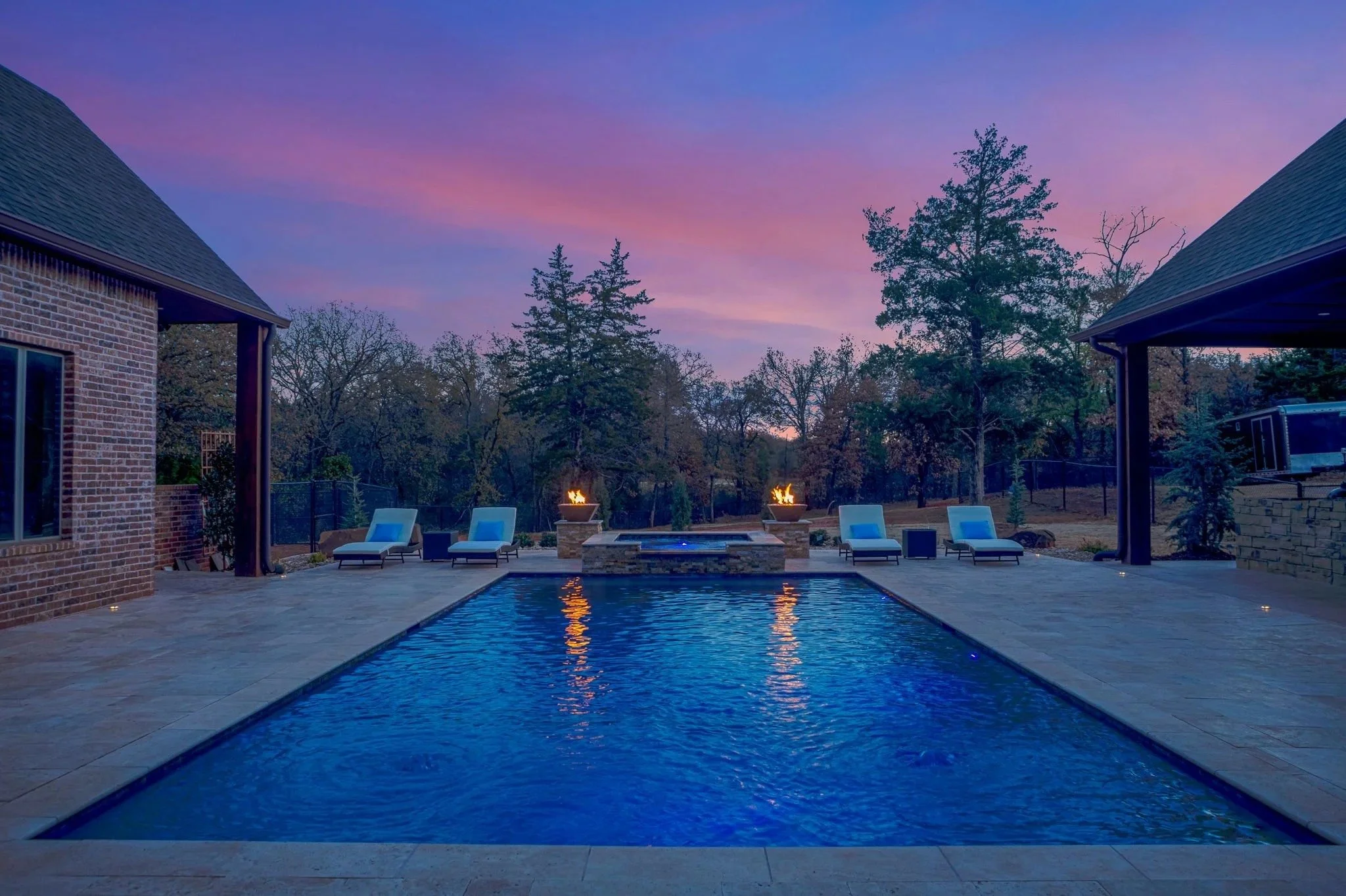 A backyard with a swimming pool at dusk, lit by fire pits, surrounded by lounge chairs, with trees and a house on either side, and a colorful pink and blue sky overhead.