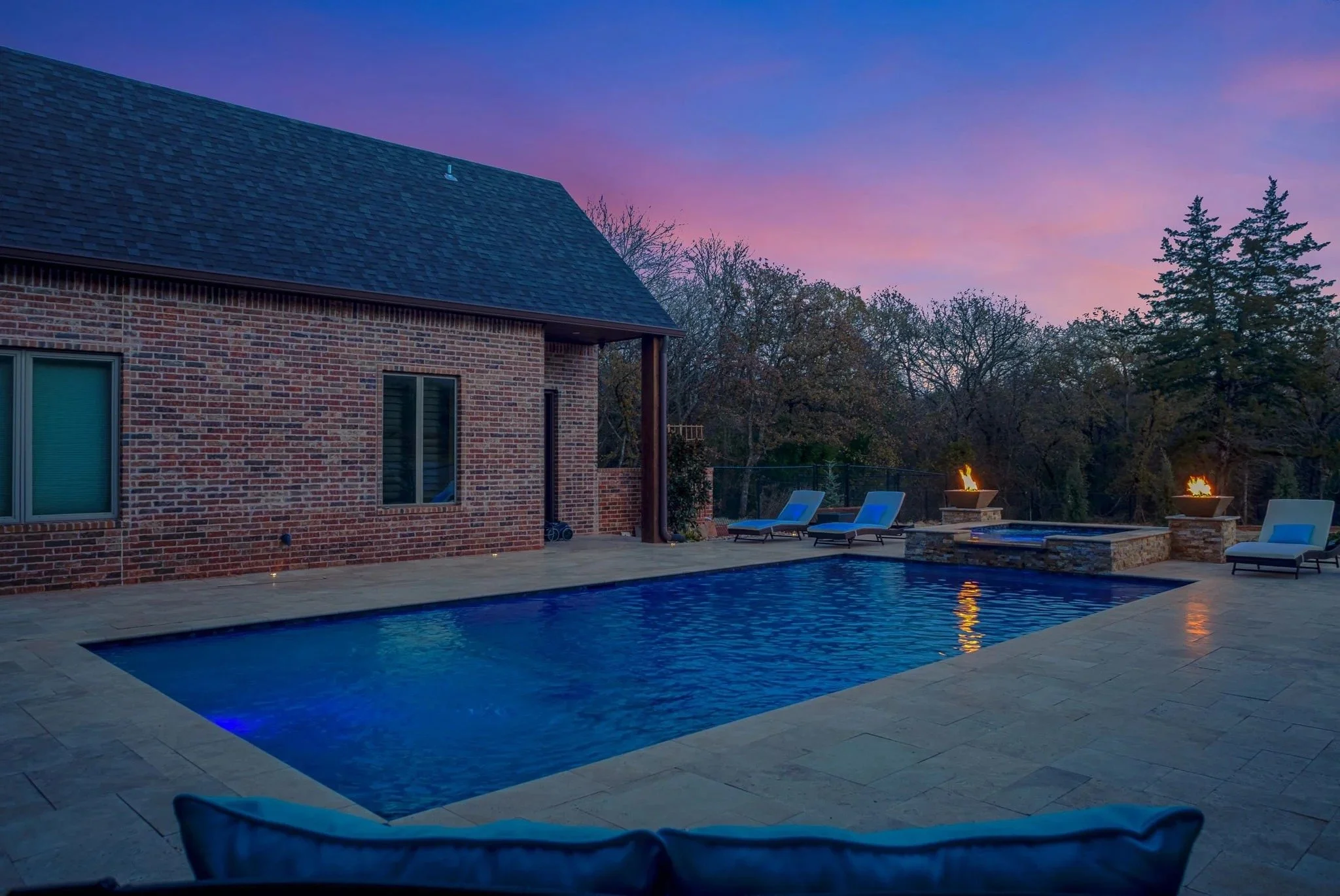 A backyard swimming pool at dusk with lounge chairs, fire bowls, a brick house wall with windows, and a sky with purple and pink hues.