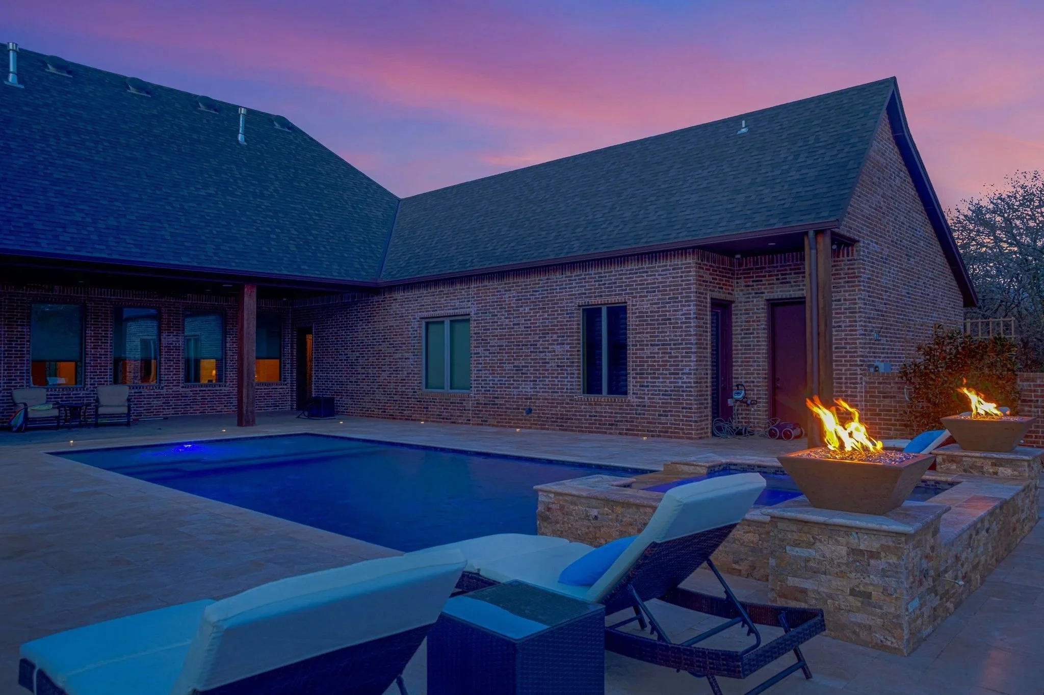 Backyard patio with swimming pool and fire pits, chairs, brick house with large windows, and a colorful sunset sky.