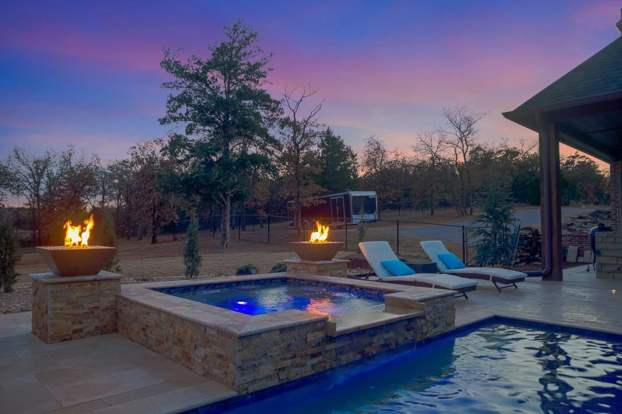 A backyard with a swimming pool, two fire pits, lounge chairs with blue cushions, trees, and a house with a roof, during sunset or early evening.