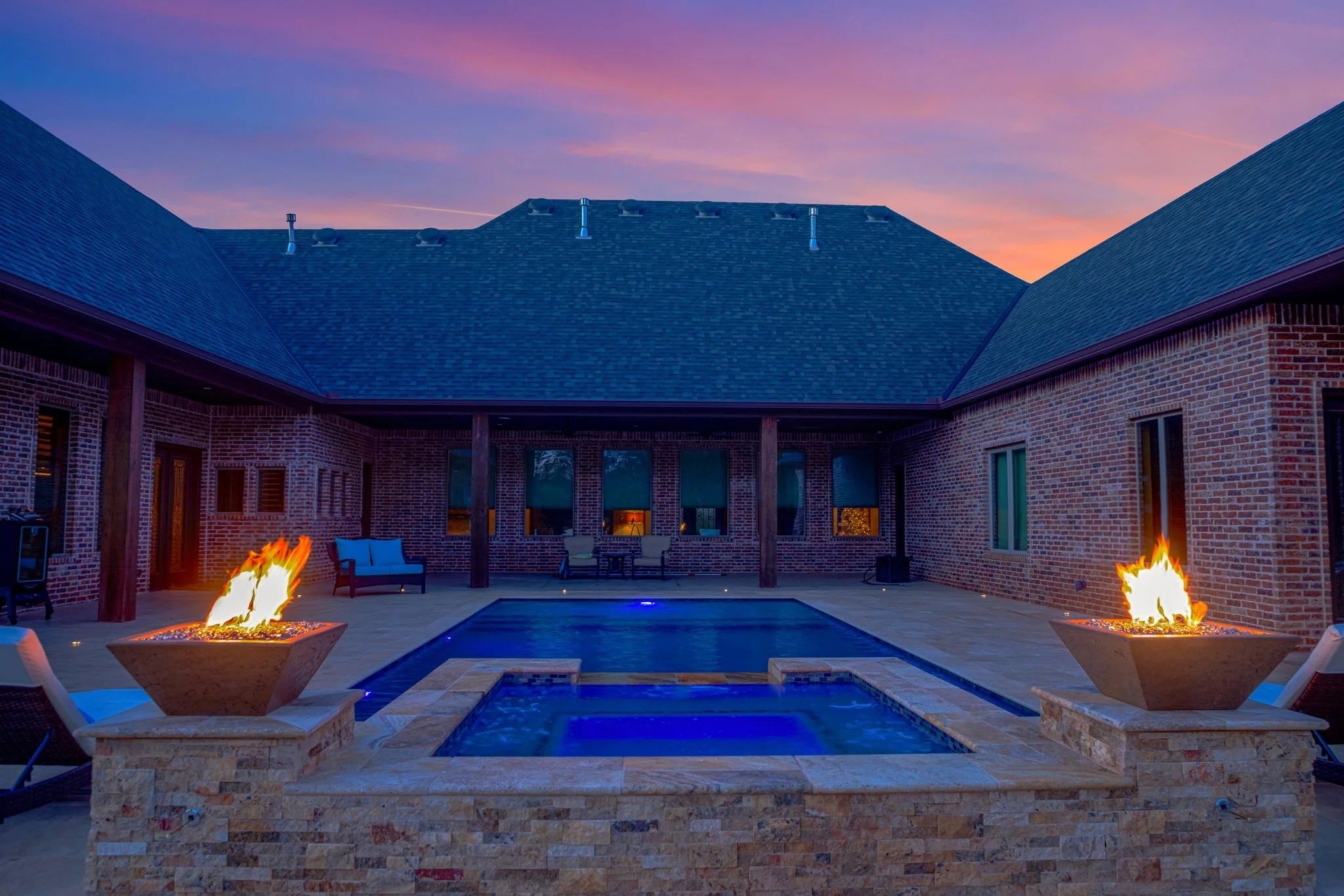 A backyard patio area featuring a swimming pool, two fire pits, outdoor seating, and a brick house with a multi-gabled roof under a colorful sky at sunset.