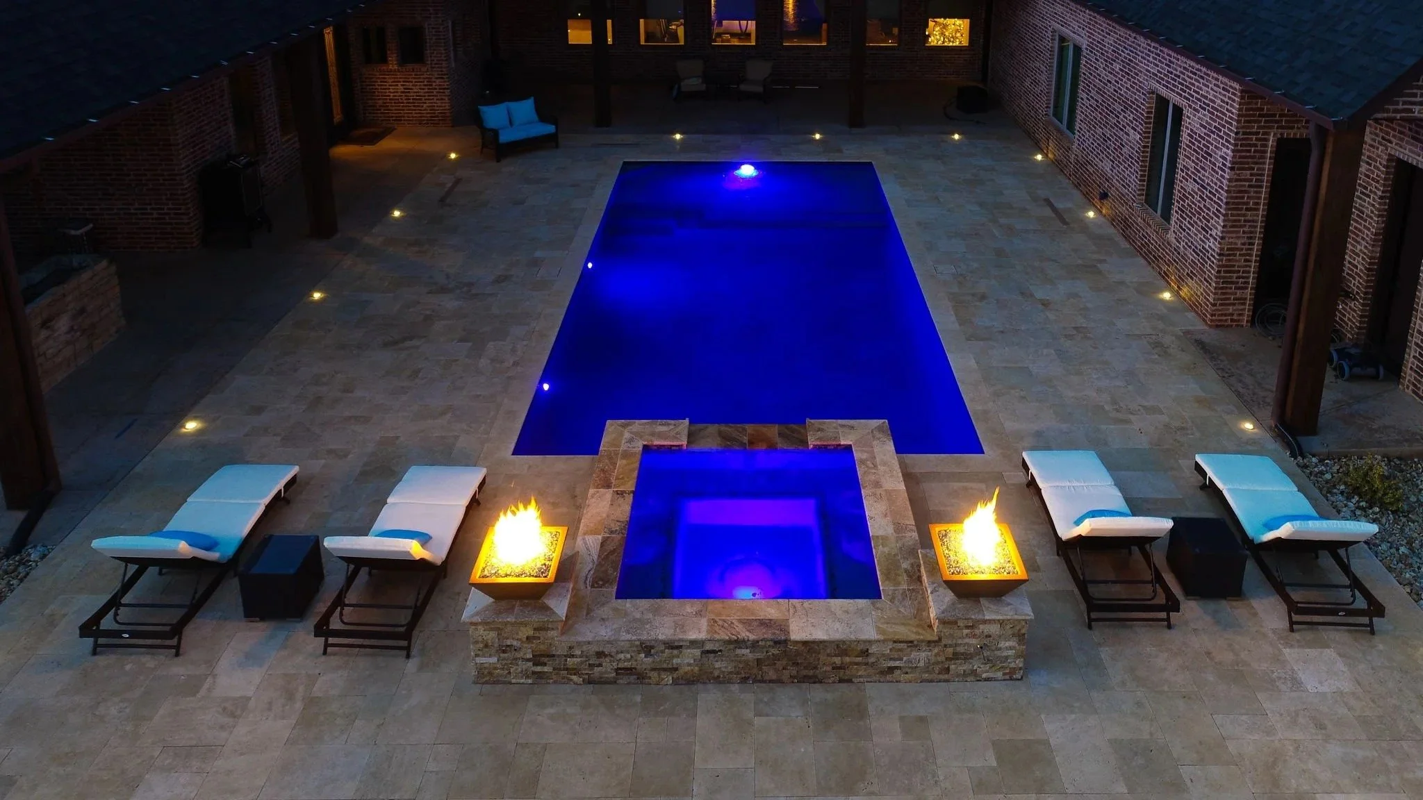 Aerial view of a backyard patio at night featuring a rectangular swimming pool with blue lighting, surrounding lounge chairs with white cushions, a fire pit with flames, and brick house walls with outdoor lighting.