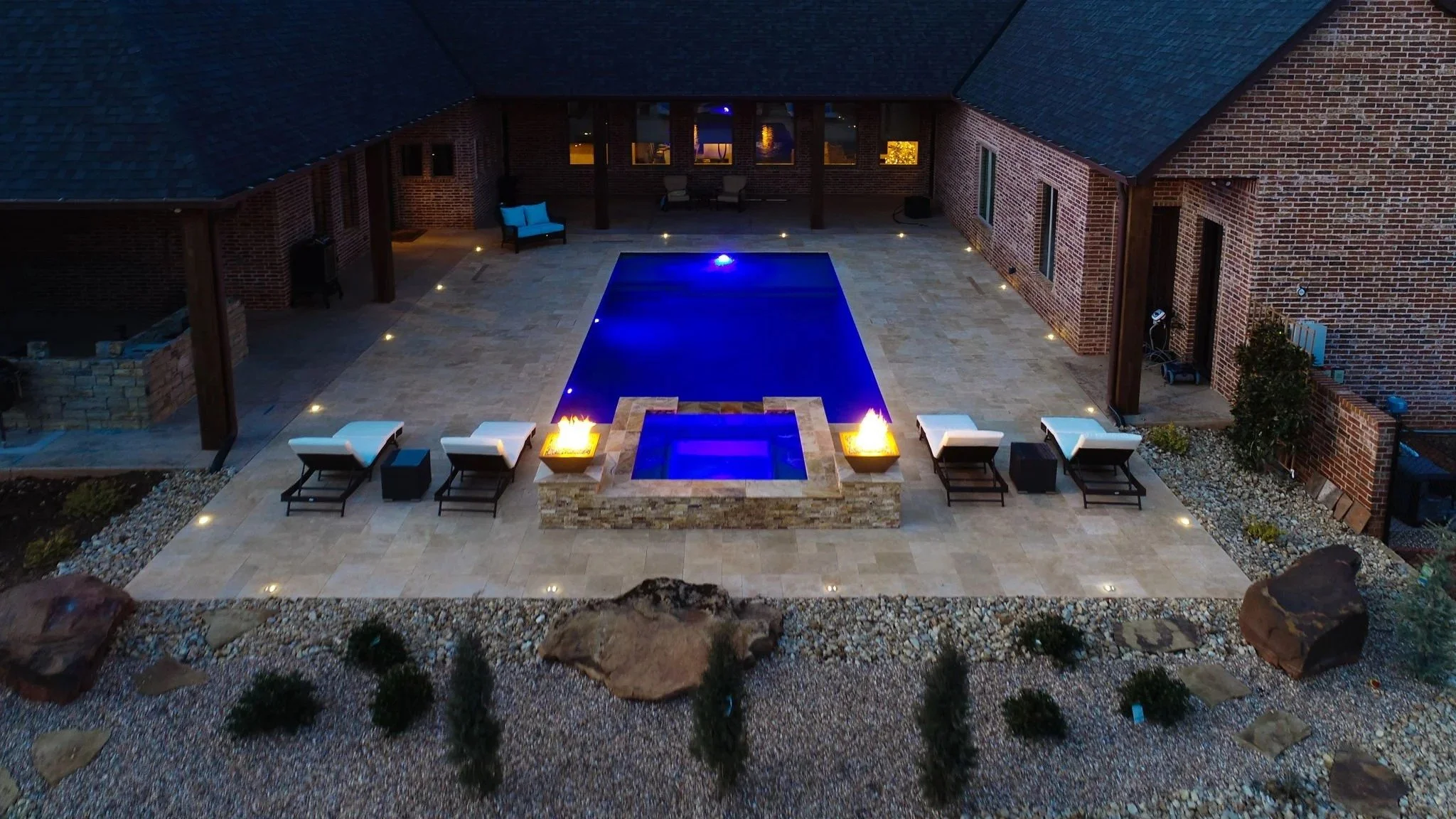 A spacious backyard with a rectangular swimming pool, illuminated by underwater lights, surrounded by lounge chairs and fire pits, with a brick house and a covered patio area in the background at night.