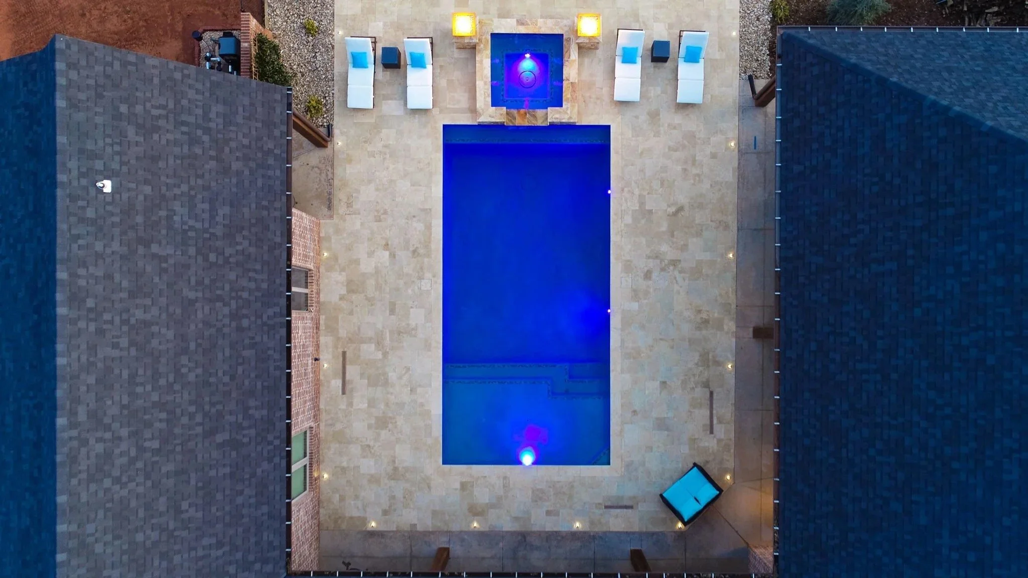 Aerial view of a backyard with a rectangular swimming pool illuminated with blue lighting, surrounded by beige stone tiles, with four white lounge chairs with blue pillows, four small square lamps, and a hot tub with blue lighting. There are two hous