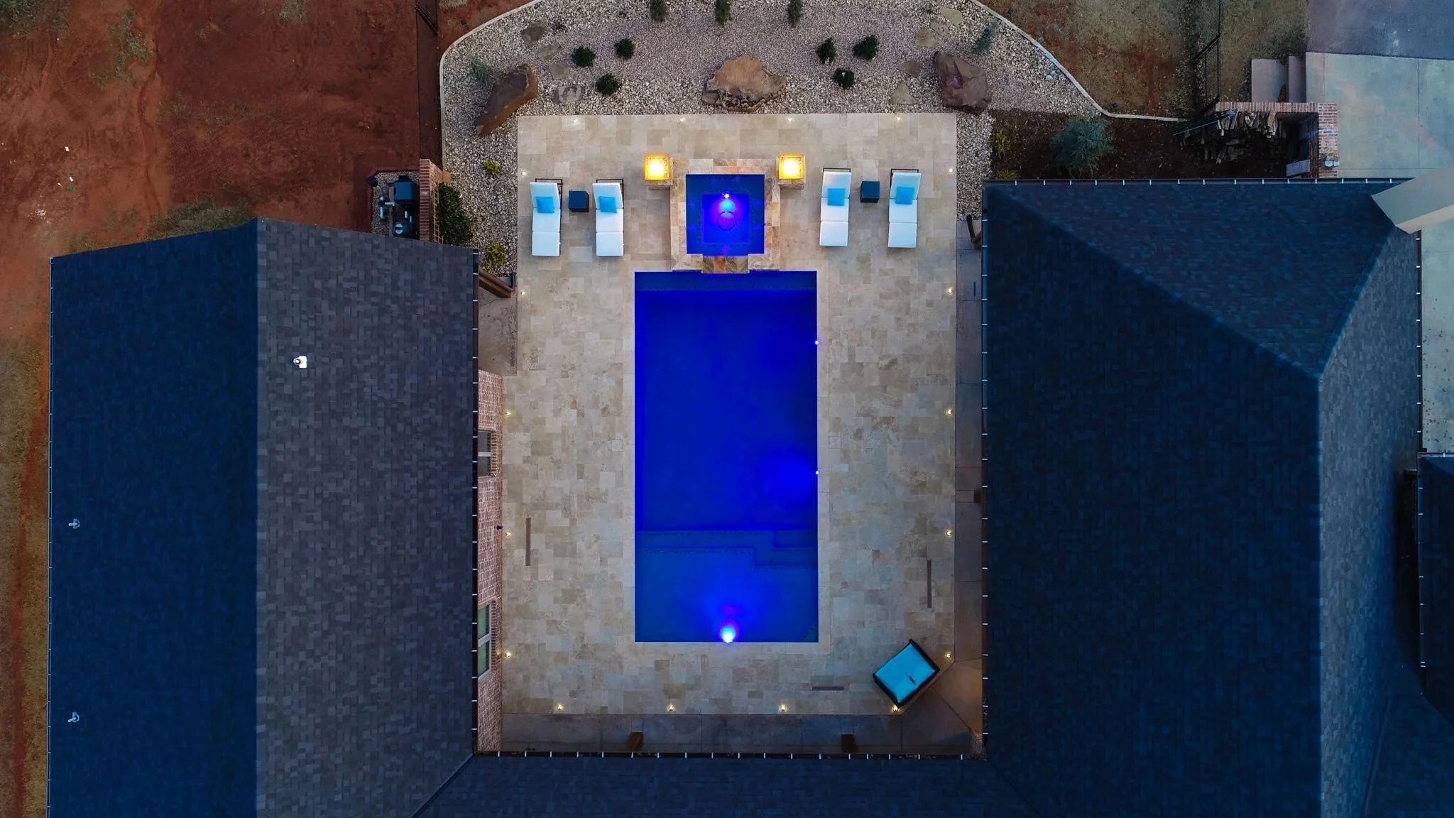 Aerial view of a backyard with a rectangular swimming pool illuminated with blue lights, surrounded by a beige tiled patio, with outdoor seating, rocks, plants, and fairy lights, bordered by neighboring houses and open land.