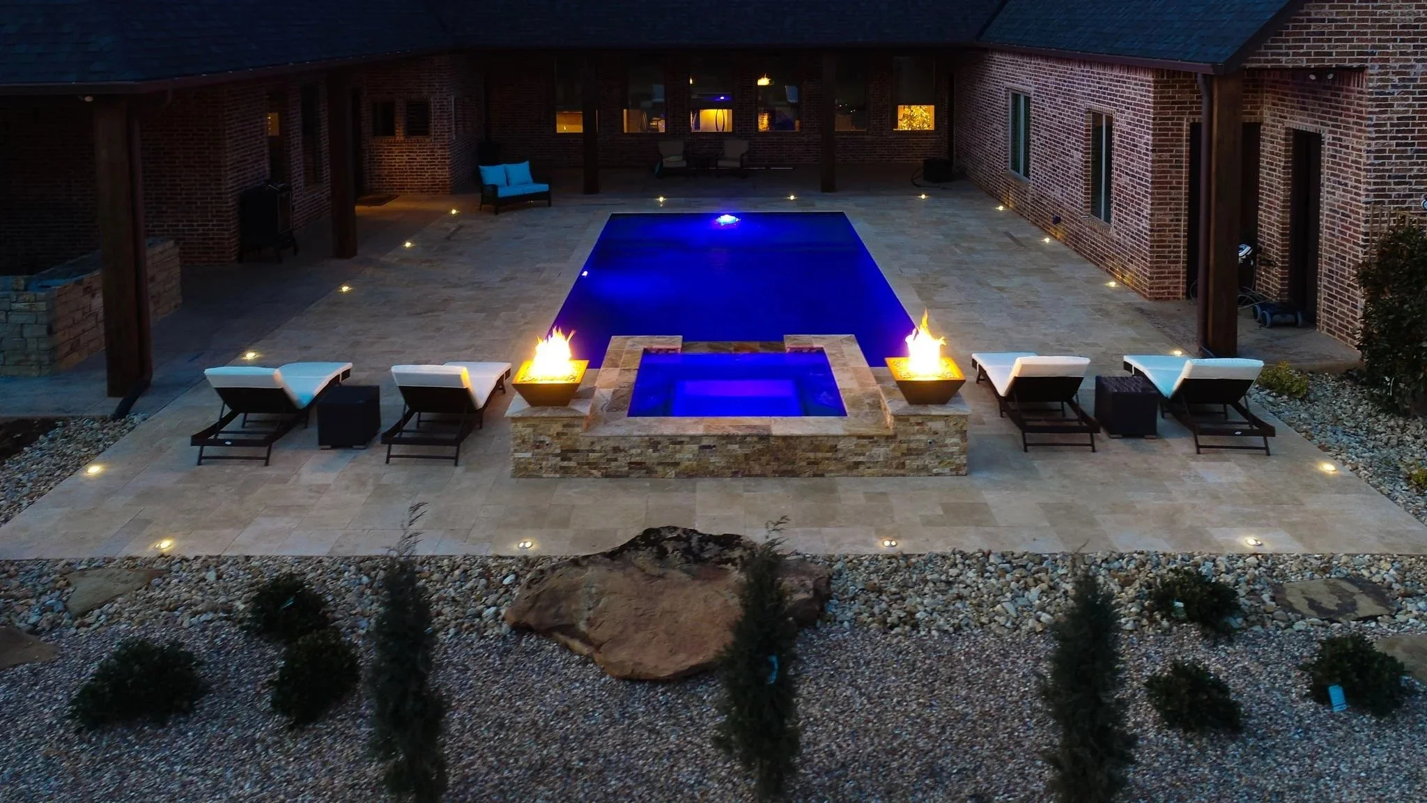Nighttime view of a backyard patio with a rectangular pool illuminated in blue, surrounded by lounge chairs, fire features, and outdoor seating, enclosed by brick walls and a covered porch.