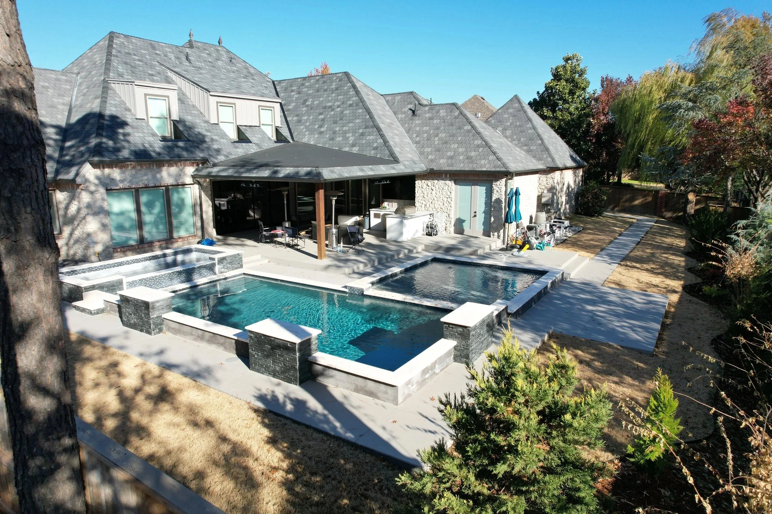 Backyard view of a house with a swimming pool, patio, and outdoor furniture, surrounded by trees and a clear blue sky.