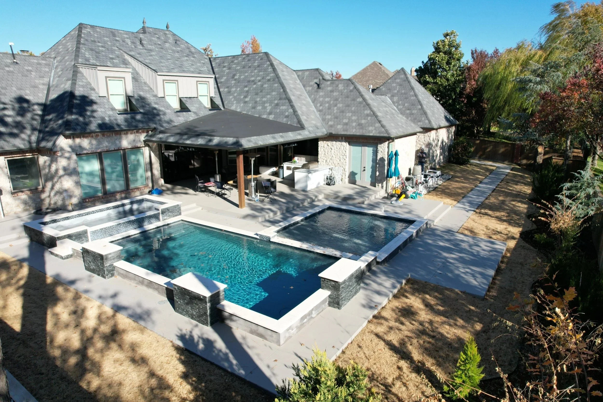 Backyard with a modern house, swimming pool, hot tub, outdoor seating, patio, and various equipment, with trees and a clear blue sky.