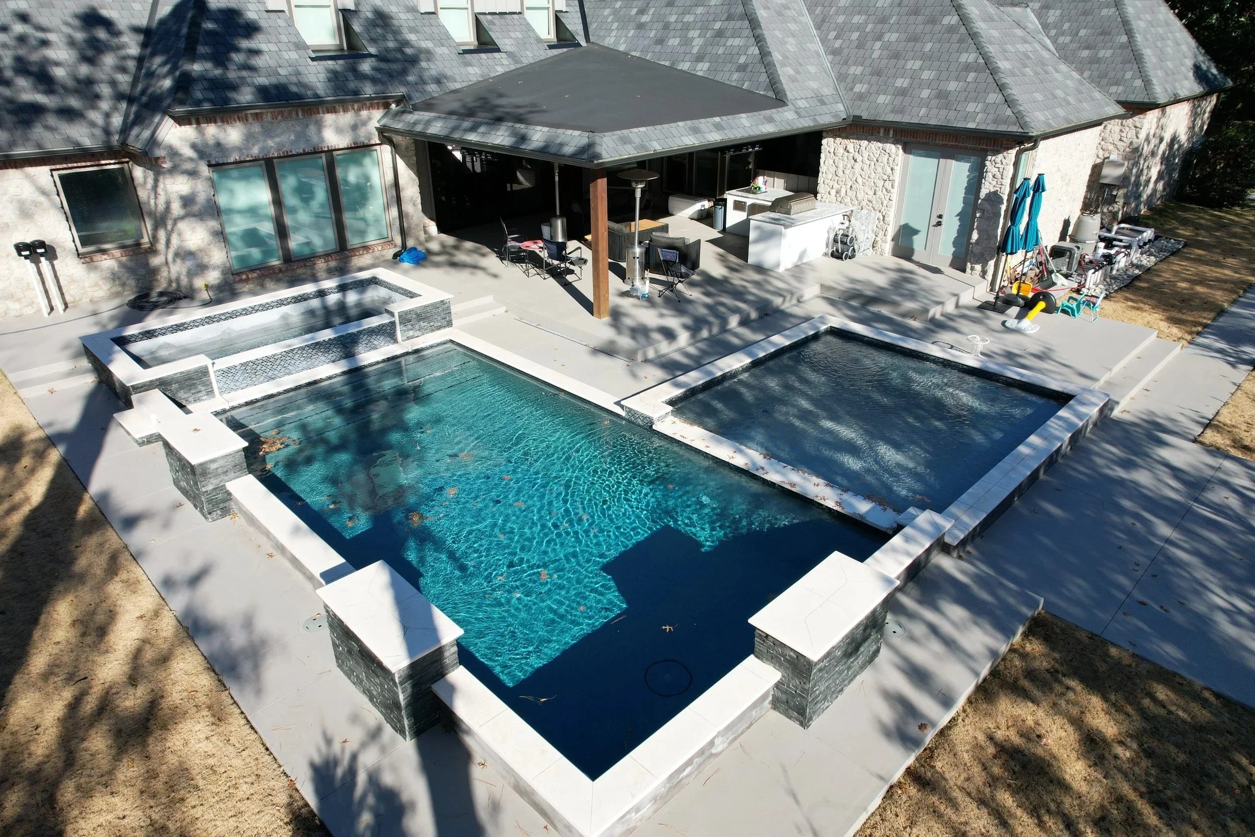 A backyard with a swimming pool and spa, with patio furniture, barbecue area, and a house with a stone facade and a slate roof.