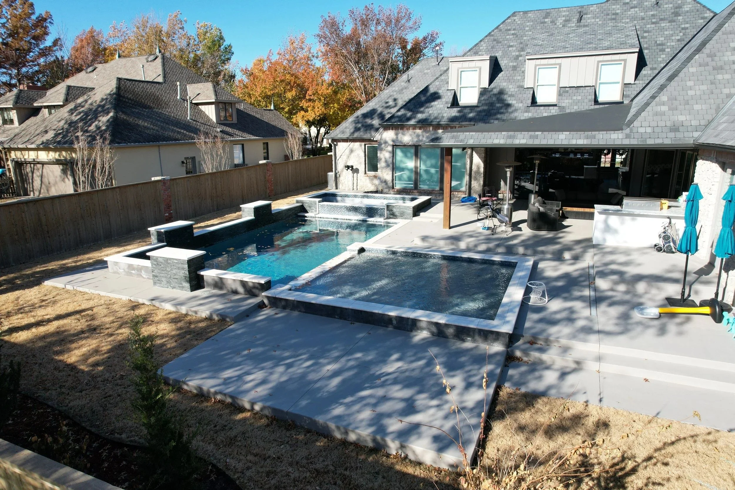 View of a backyard with a swimming pool, concrete patio, nearby house with gray shingle roof, and trees with autumn foliage in the background.