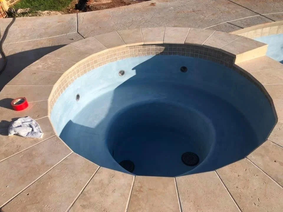Empty round hot tub with light blue interior tiles, surrounded by beige stone tiles, outside on a patio.