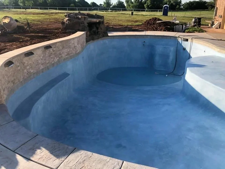 Empty swimming pool with a blue interior, surrounded by a stone and concrete deck, in a backyard with construction or renovation work in progress.