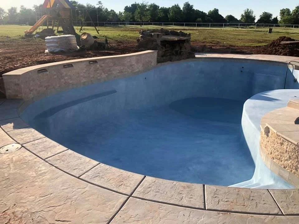 Empty in-ground swimming pool under construction in a backyard with a slide and a grassy field in the background.
