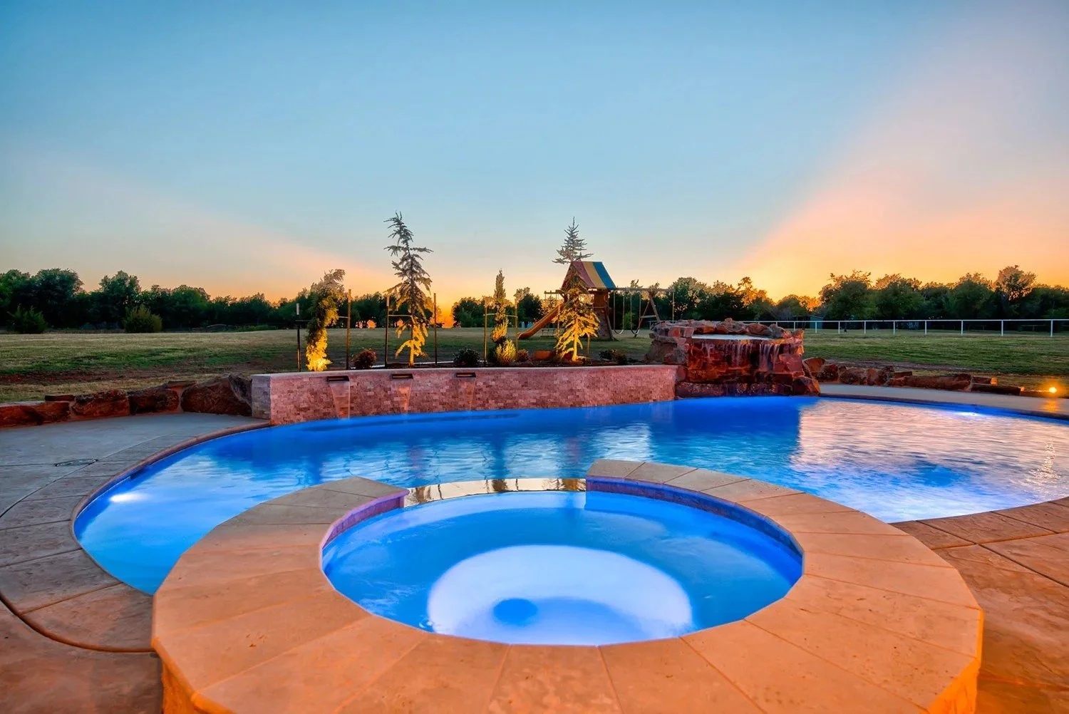 An outdoor swimming pool with illuminated hot tub at sunset, with a grassy field, trees, and playground in the background.