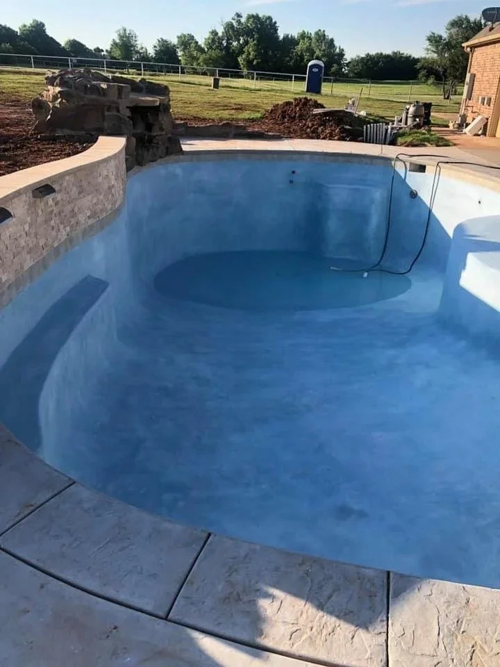 Empty swimming pool in backyard under clear sky with trees and grass in the background.