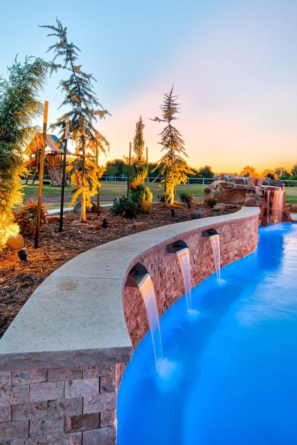 A backyard with a curved stone and brick water fountain with three spouts, water flowing into a blue pool, tall trees, a playground structure in the background, and a sunset sky.