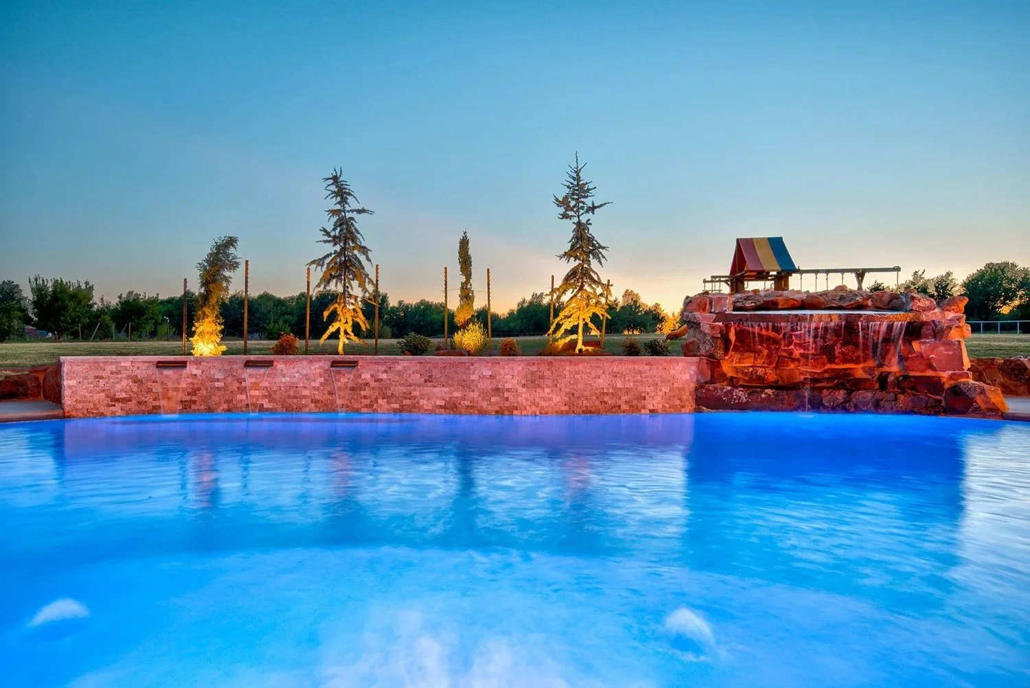 A swimming pool with a waterfall feature at sunset, surrounded by grass and trees, with a small colorful cabana on the rocks.