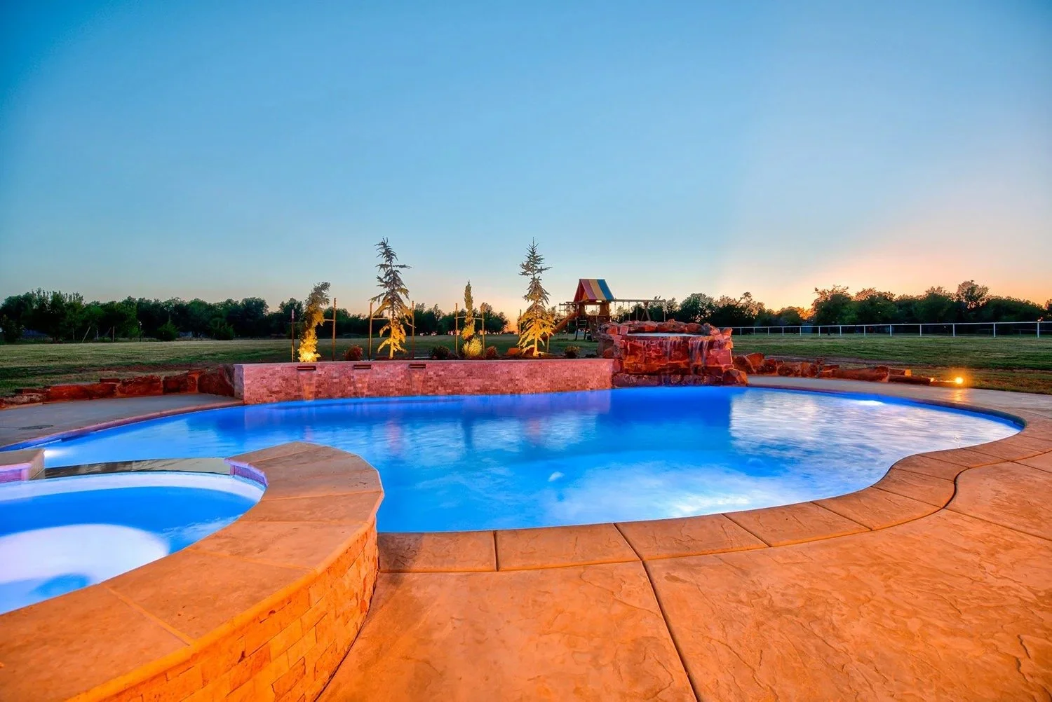 An outdoor swimming pool with a hot tub next to it, surrounded by a paved patio. There are decorative trees and a small play area with a slide in the background, against a sunset sky.