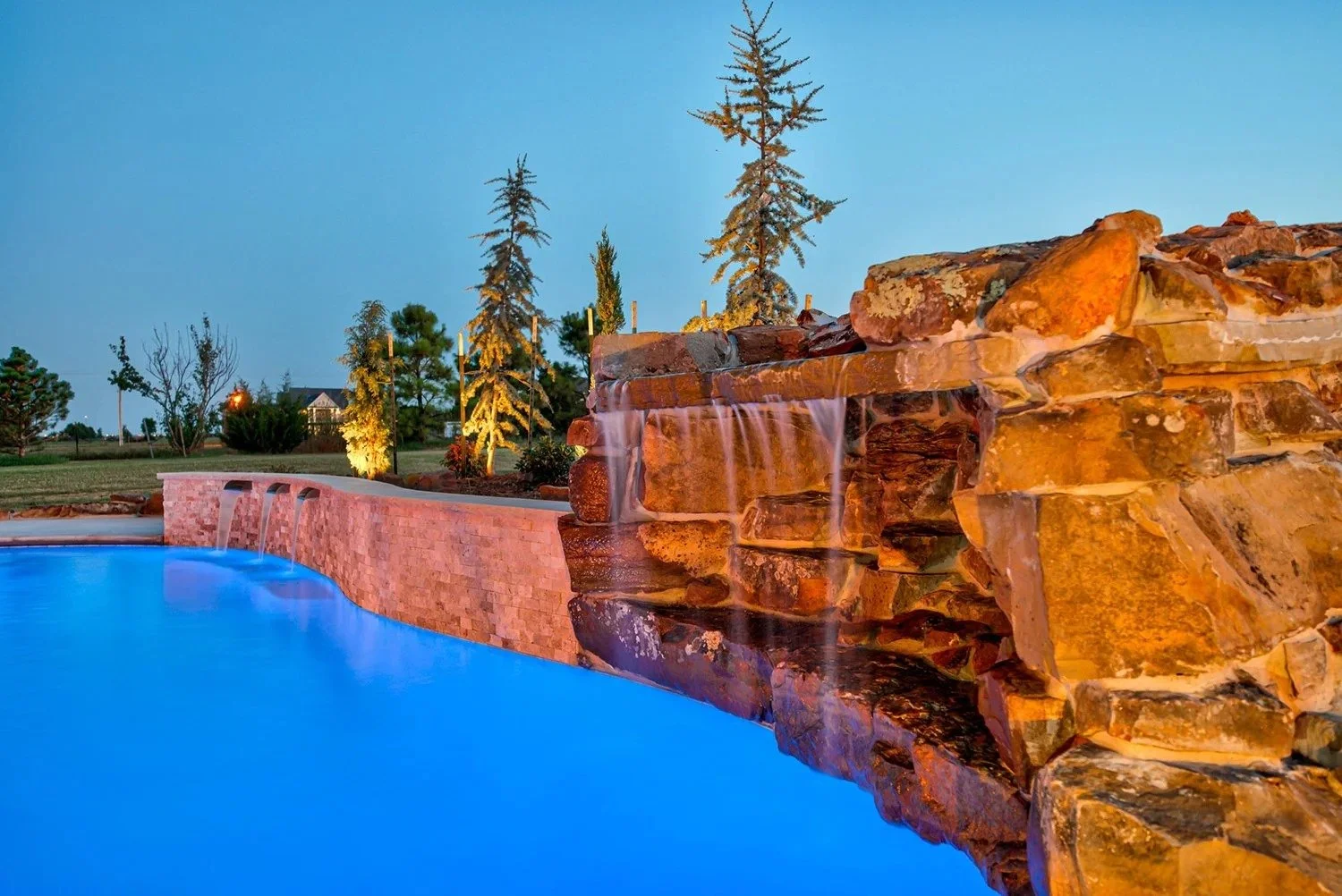 An outdoor swimming pool with a stone waterfall feature at dusk, surrounded by trees and landscaped grounds.