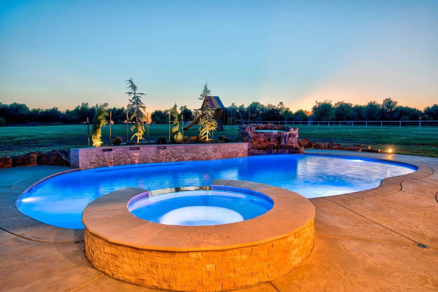 A backyard with a lit swimming pool and hot tub at dusk, surrounded by a stone deck, trees, and a grassy field.