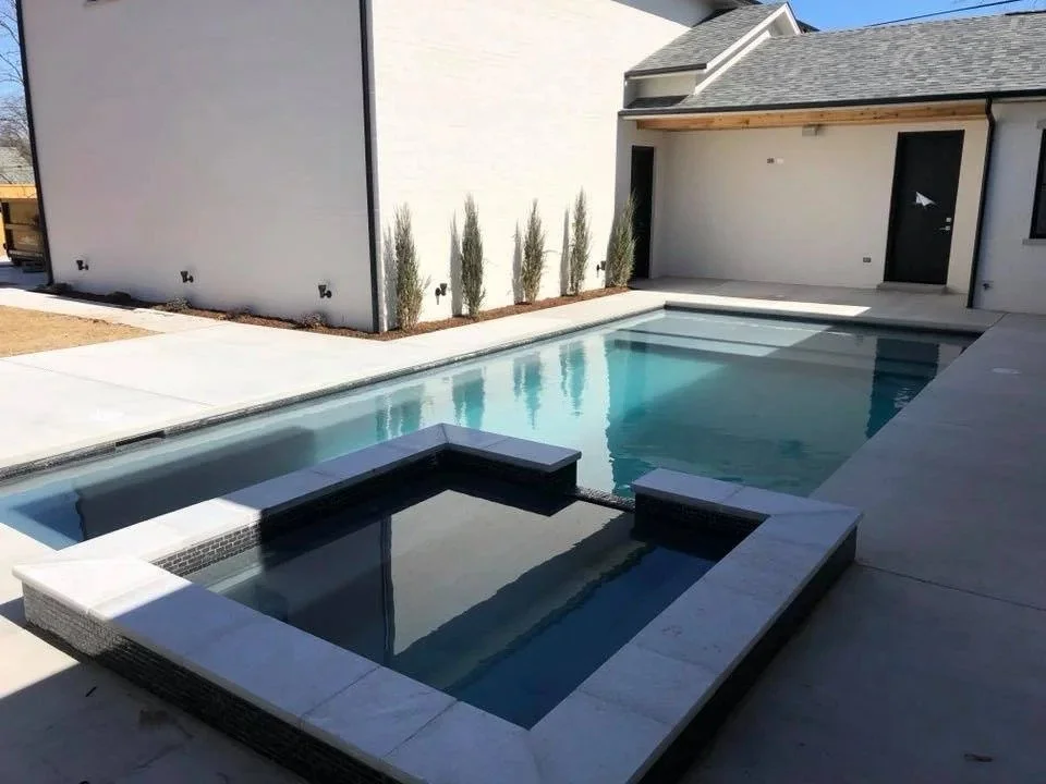 Backyard with a modern swimming pool and hot tub, white house wall with small trees, concrete pool deck, and patio door.