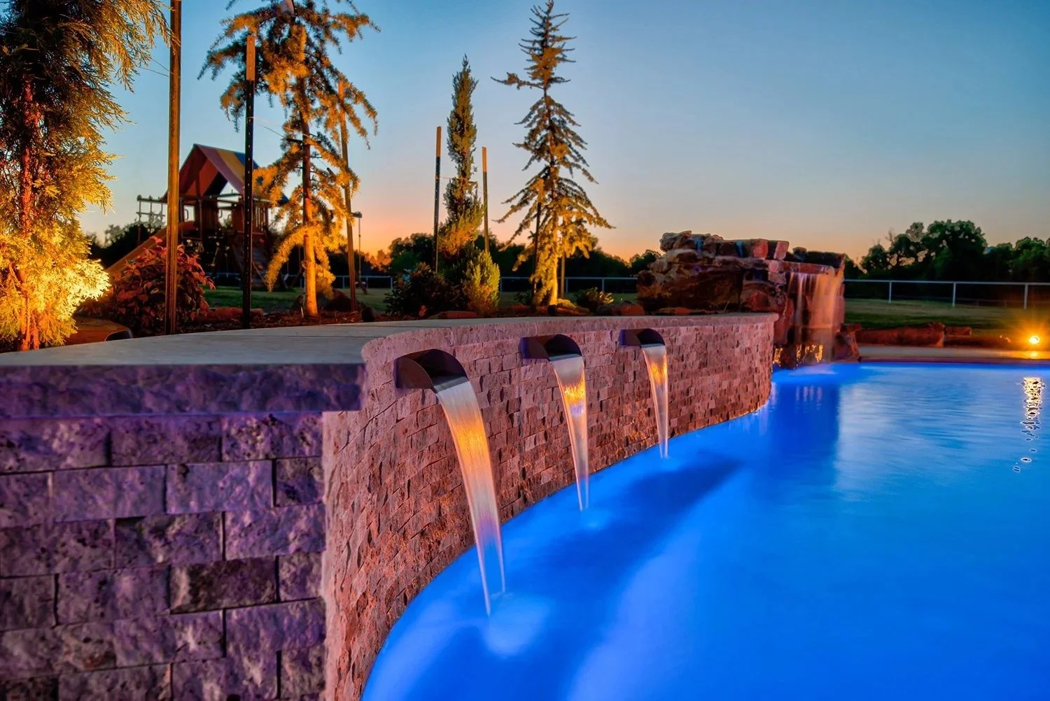 A swimming pool with blue water illuminated by underwater lights, a brick fountain with three water spouts, and a park with trees and playground equipment in the background during sunset.